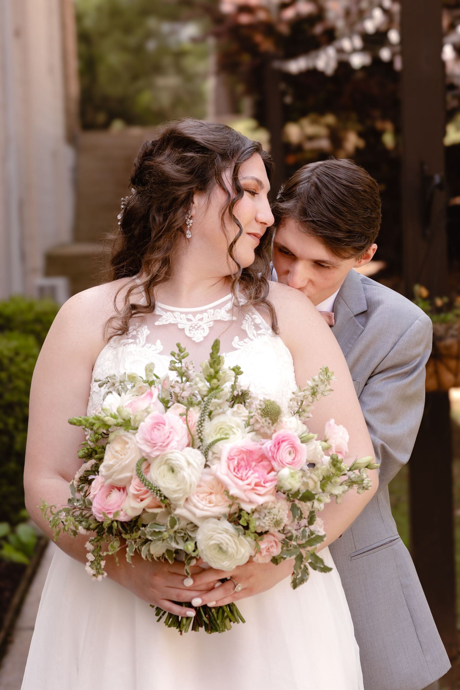 Bride in white lace gown holds pink and white bouquet as groom in gray suit nuzzles her neck outdoors.
