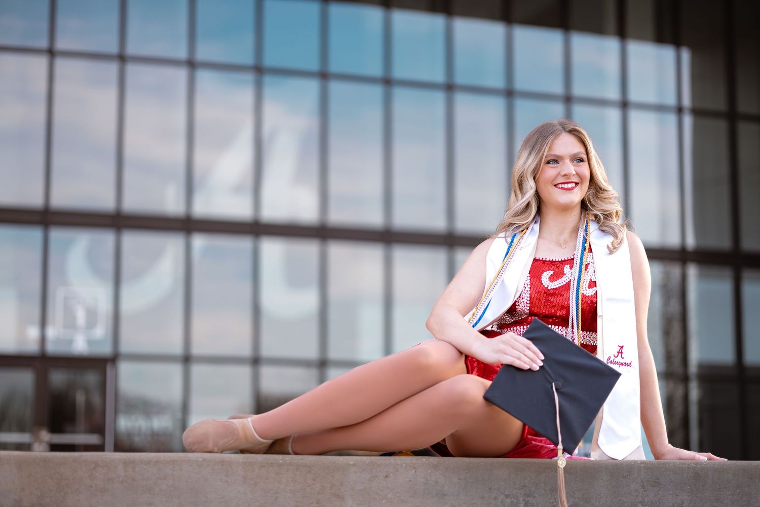 Smiling female graduate in red and white regalia sitting outdoors holding cap during graduation photo session.