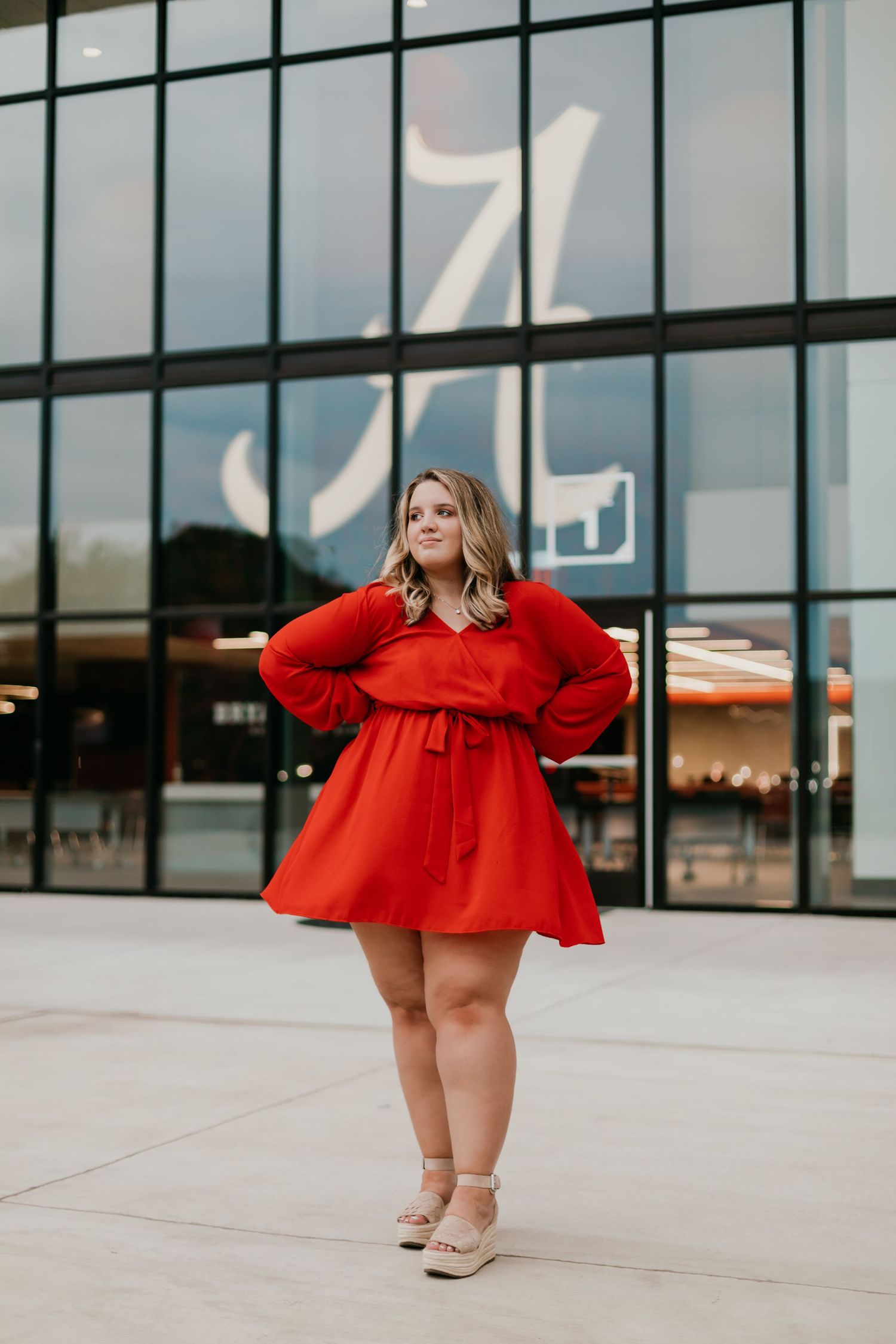 Woman in vibrant red flowy dress poses confidently outside modern building with glass facade on sunny day.