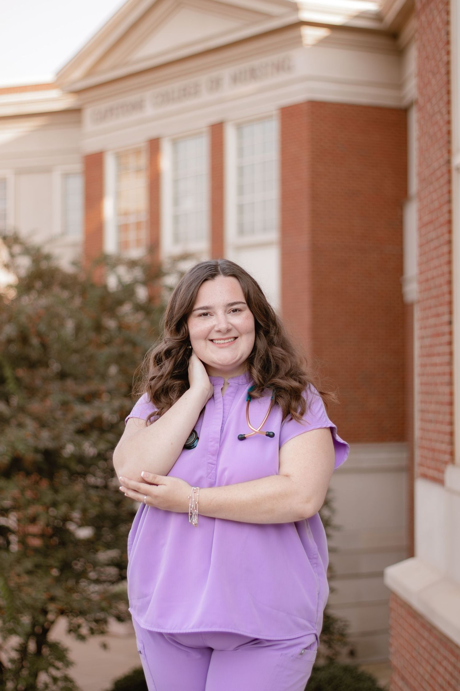 A person in purple scrubs stands smiling with crossed arms in front of a brick building.