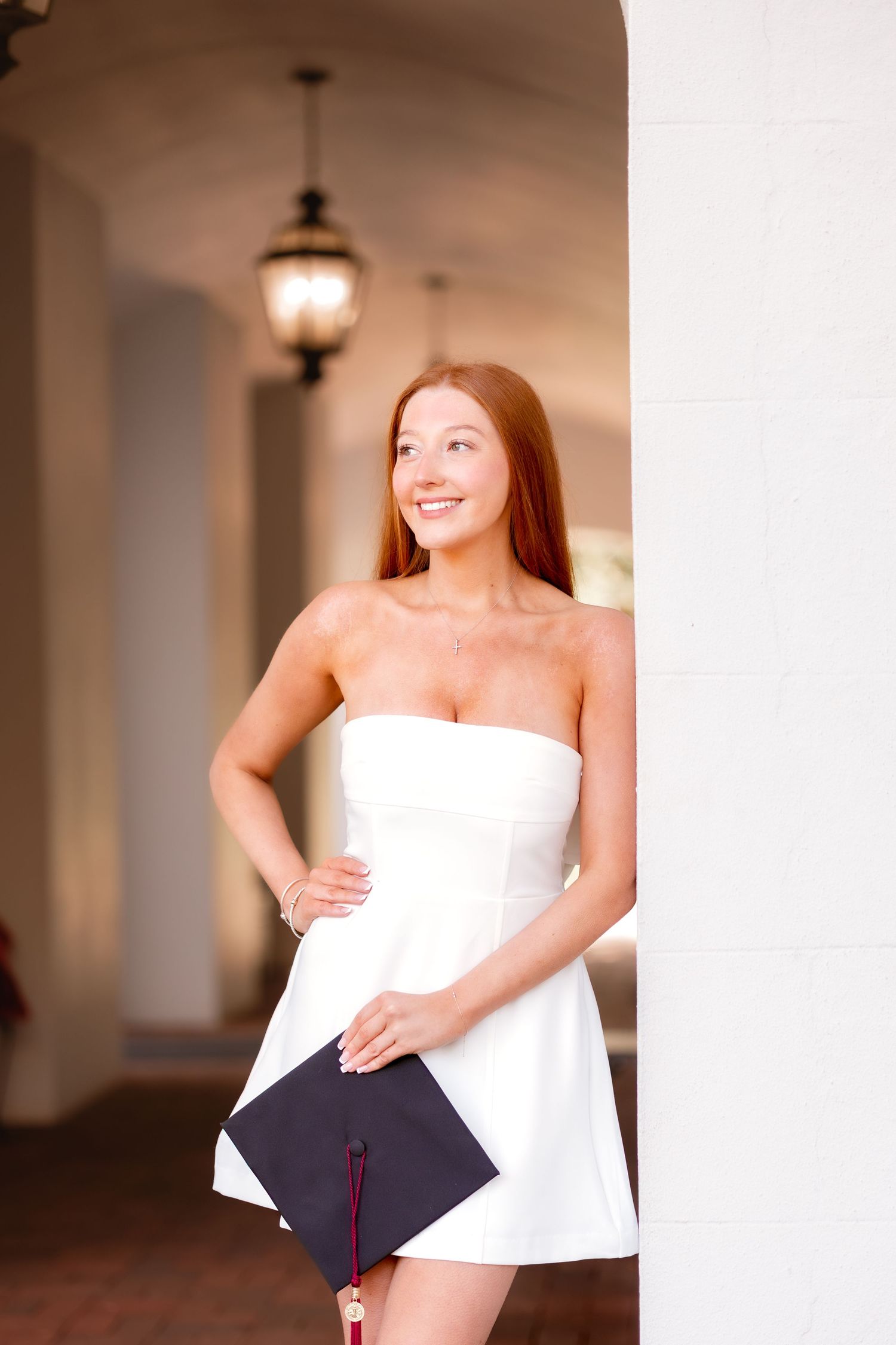 A person in a white graduation dress holds a cap while standing in an arched hallway.