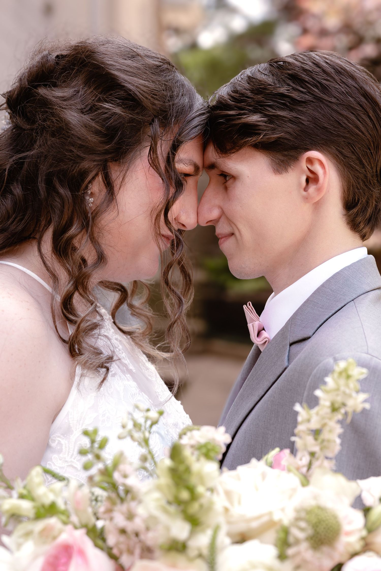 Bride and groom touching foreheads romantically, surrounded by pink and white wedding flowers.