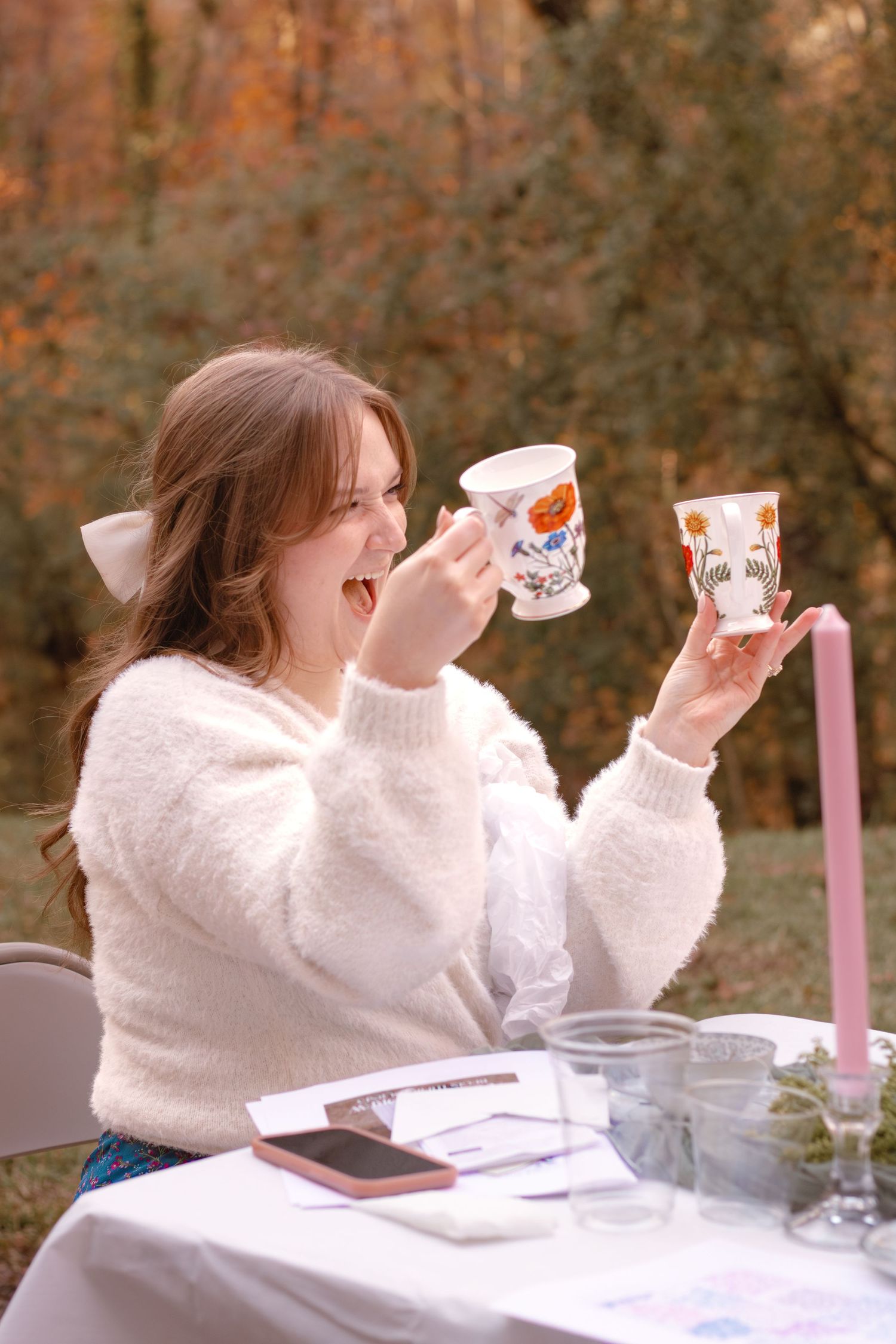 Person in white sweater laughing while holding tea cups at an outdoor table setting with autumn foliage in background.