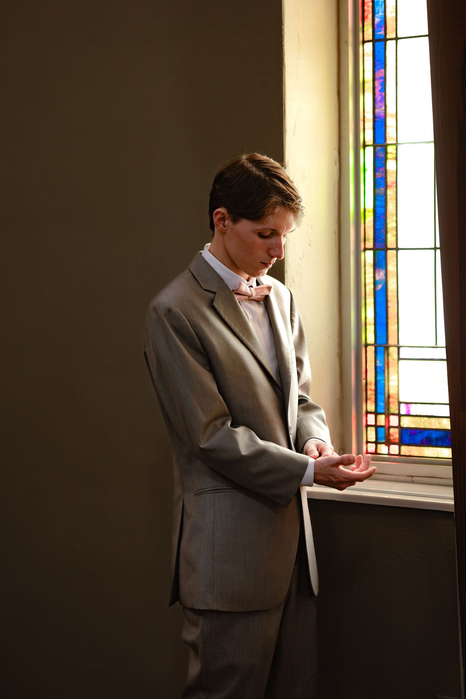 Young man in gray suit and bow tie standing by stained glass window in church, looking down thoughtfully.