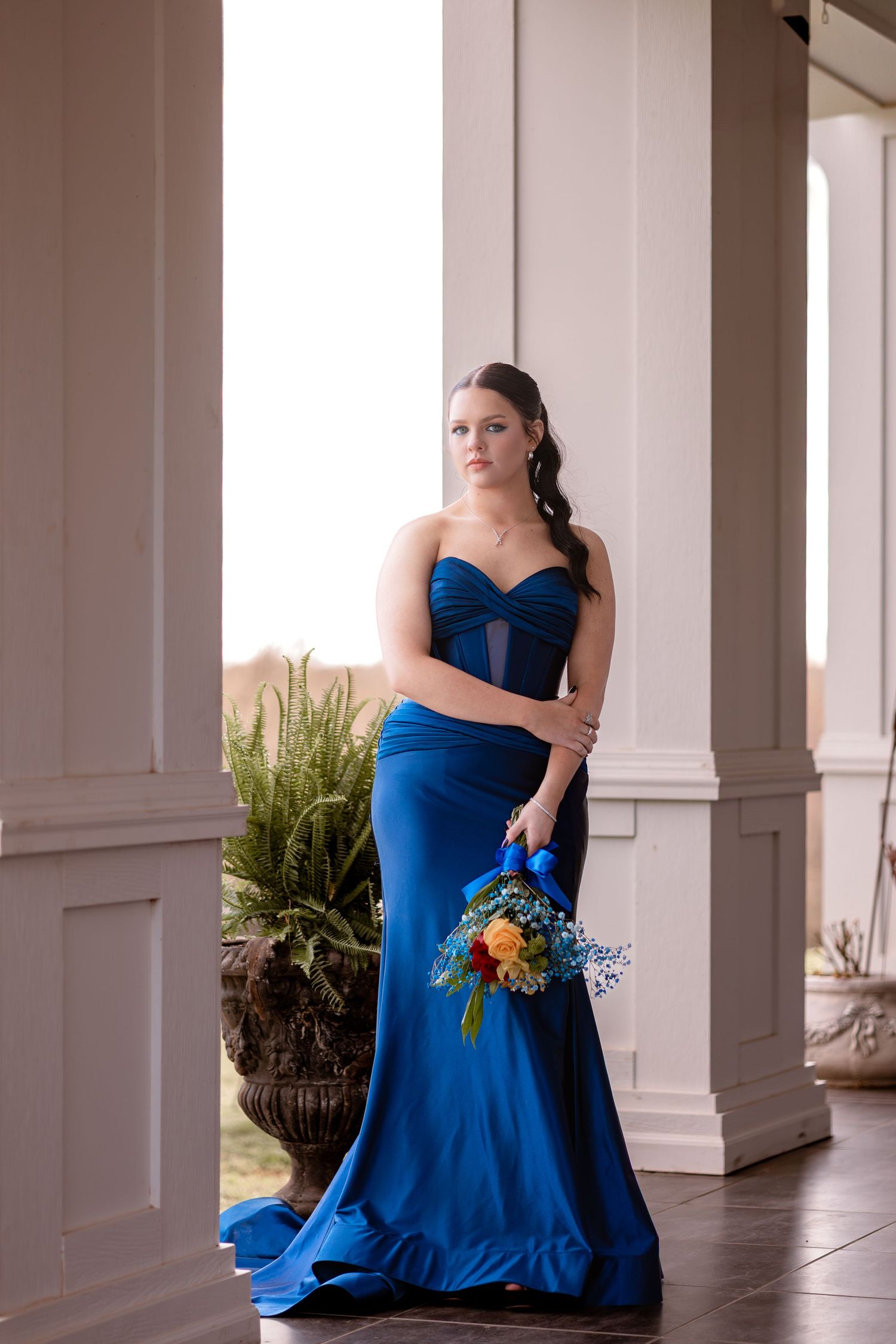 A person in a royal blue formal gown holds a colorful bouquet while posing on an elegant porch.