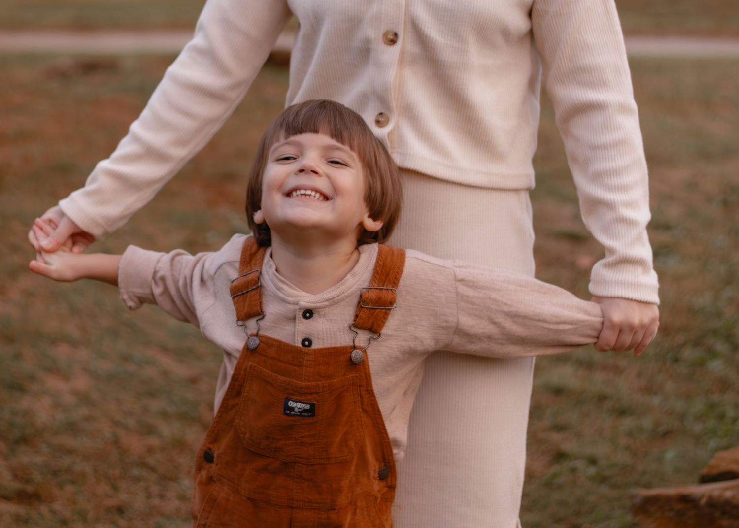 A child in brown overalls plays joyfully outdoors with an adult in beige clothing.