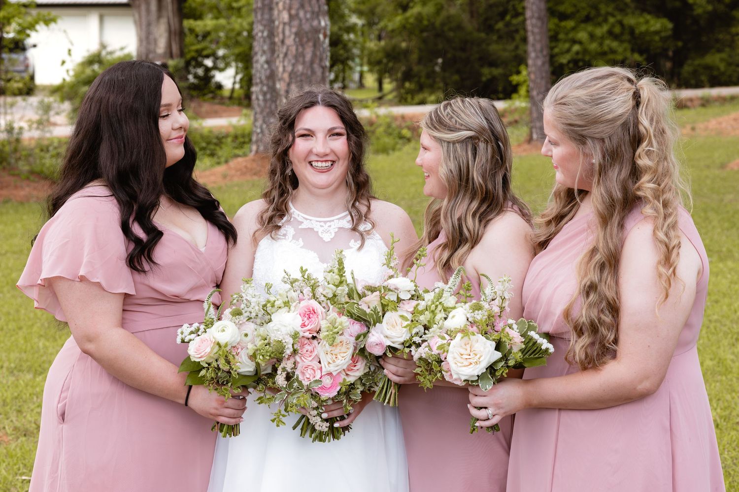 Bride in white gown holds floral bouquet with three bridesmaids in pink dresses outdoors at a wedding.