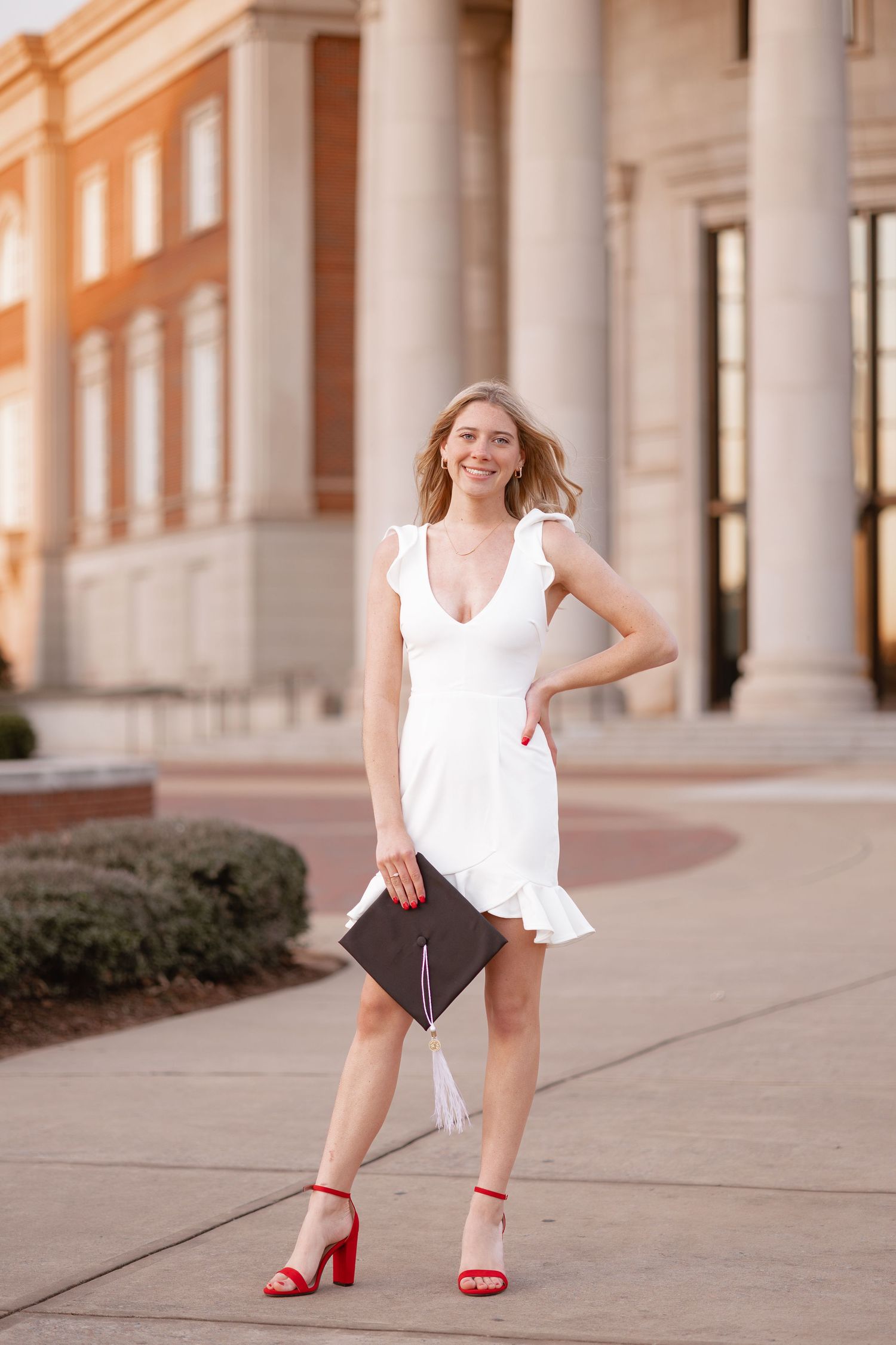 Graduate in white dress and red heels poses against columned architecture.