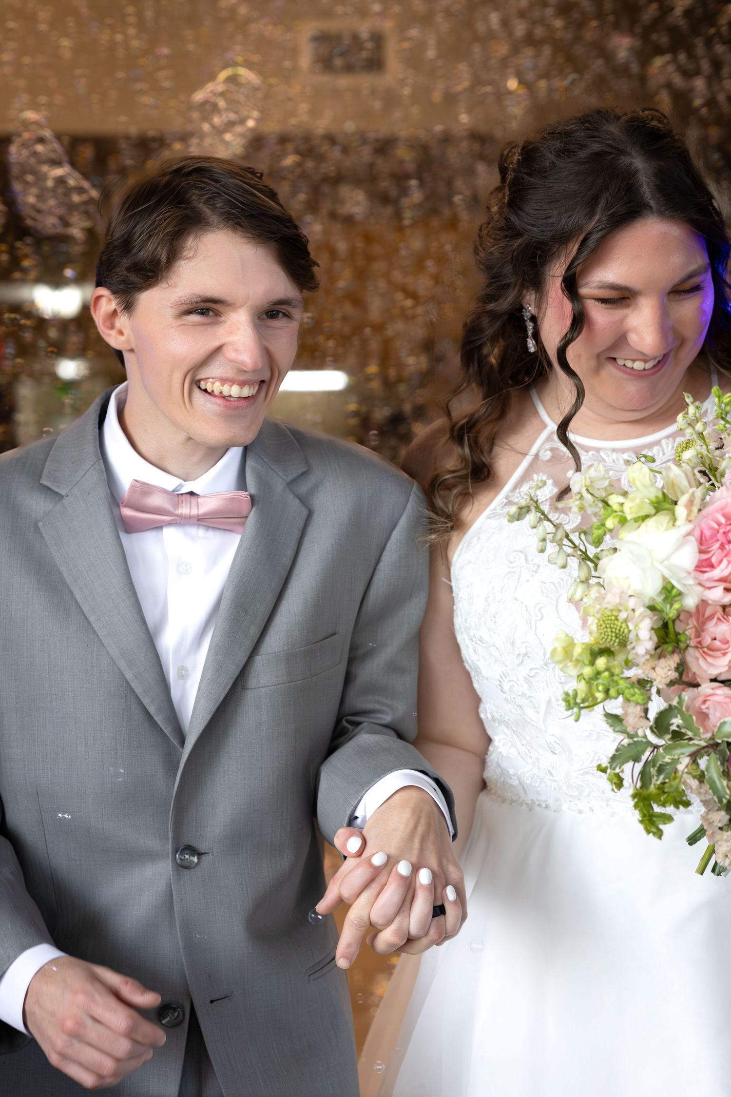 Smiling newlyweds holding hands, groom in gray suit with pink bow tie, bride in white gown holding pink bouquet.