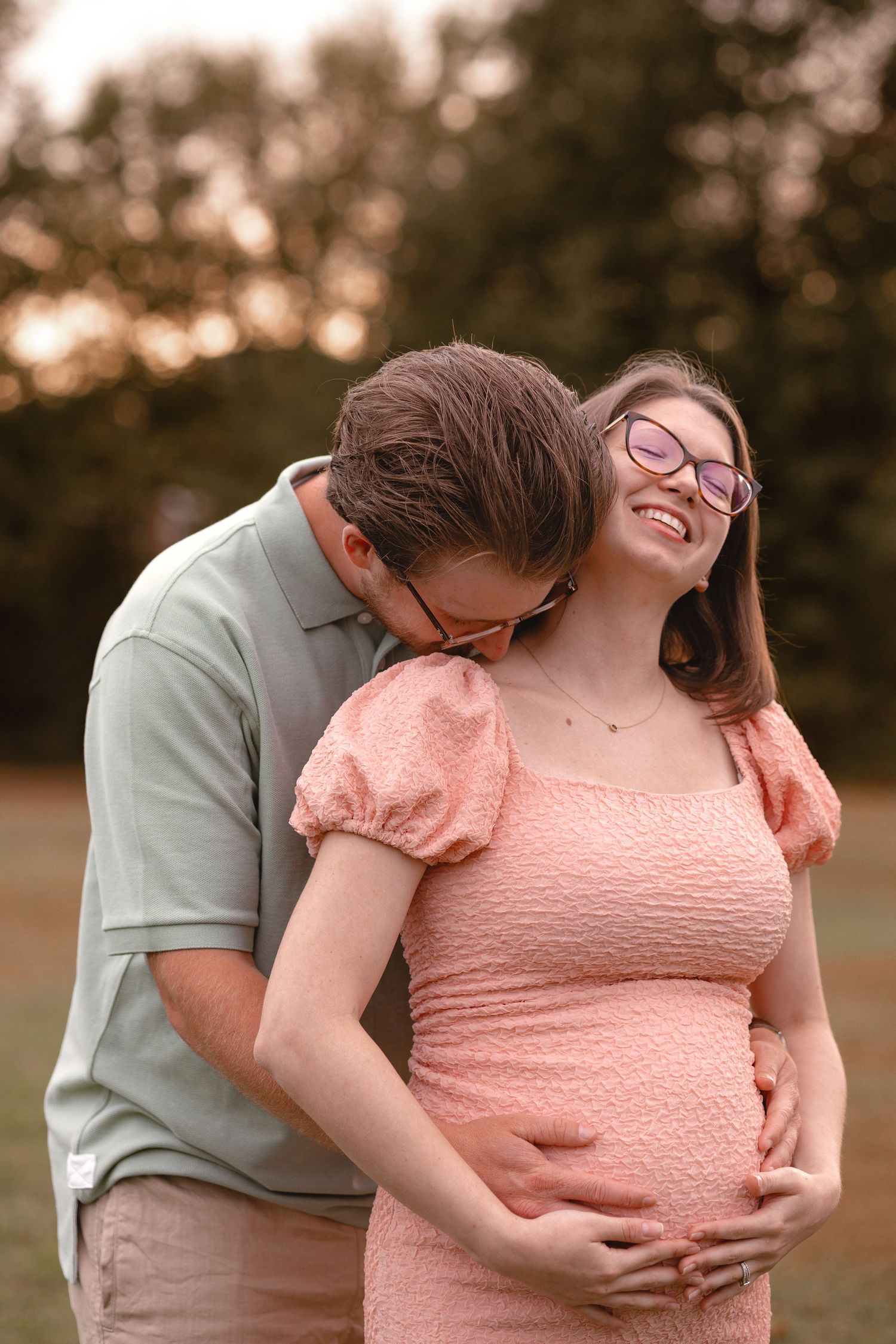Happy pregnant woman in pink dress with partner kissing her cheek outdoors at sunset.