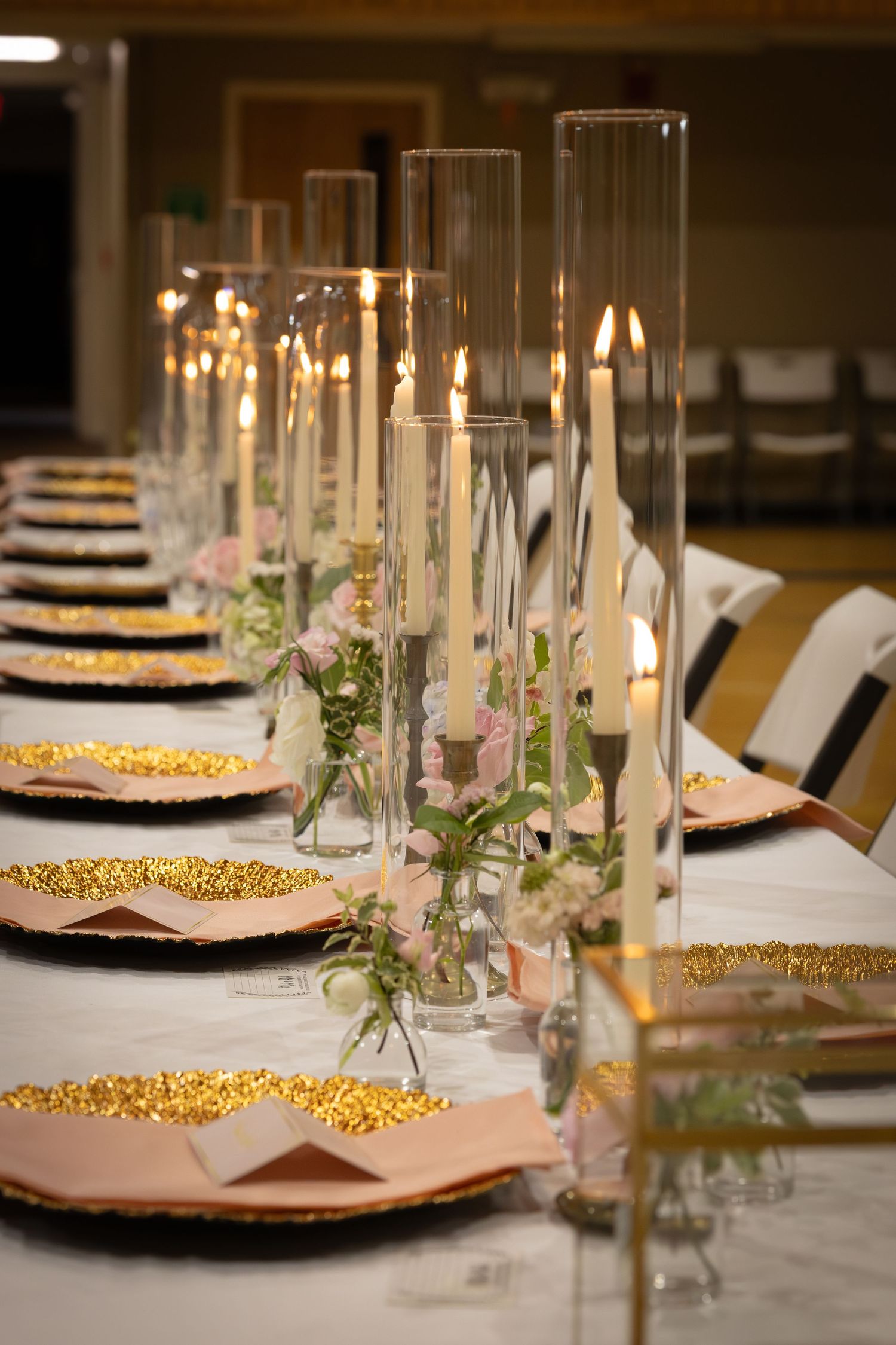 Elegant wedding reception table with tall glass candle holders, gold glitter charger plates, and delicate floral arrangements.