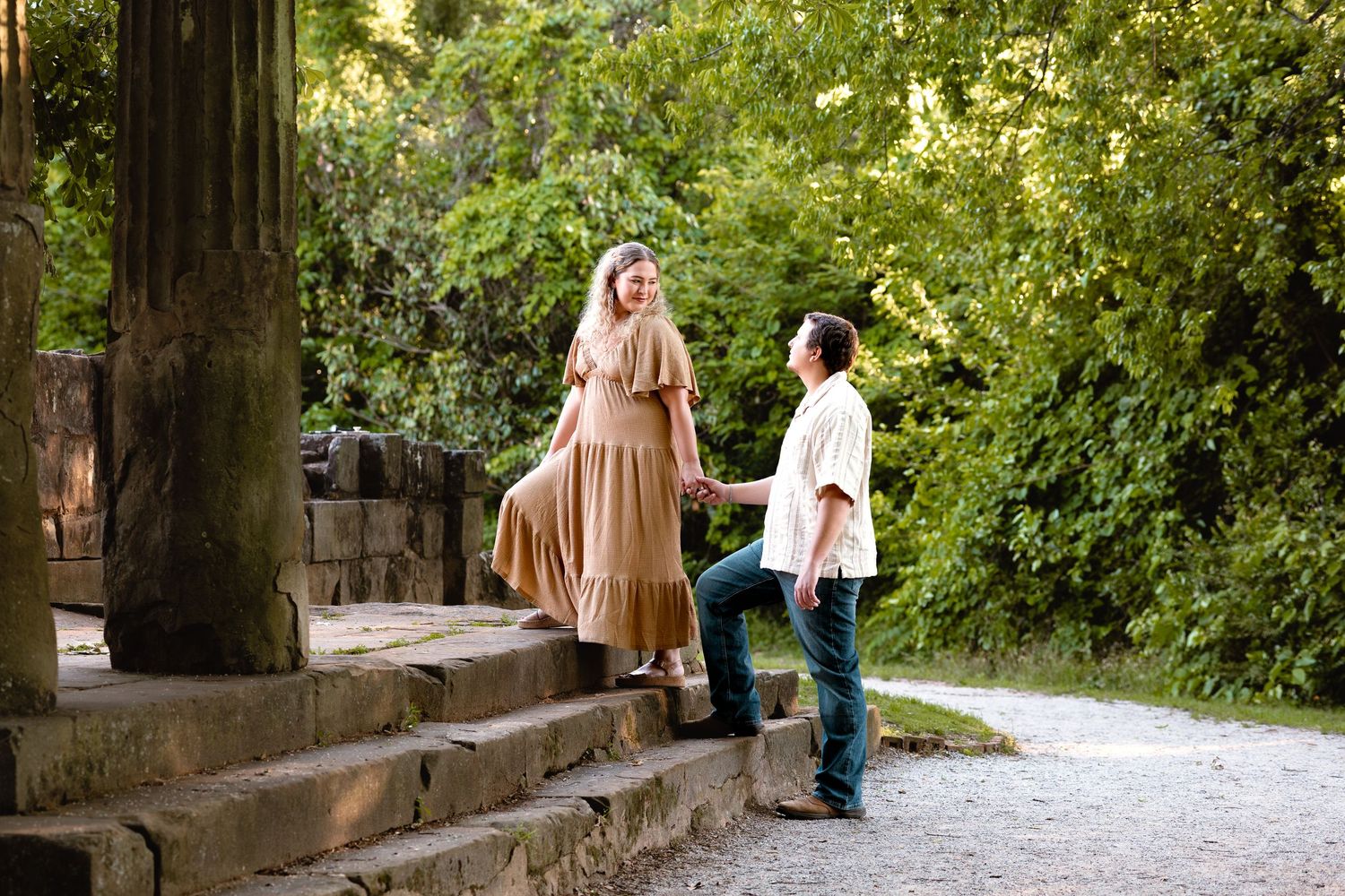 Couple holding hands on stone steps surrounded by lush green trees in a romantic outdoor setting.