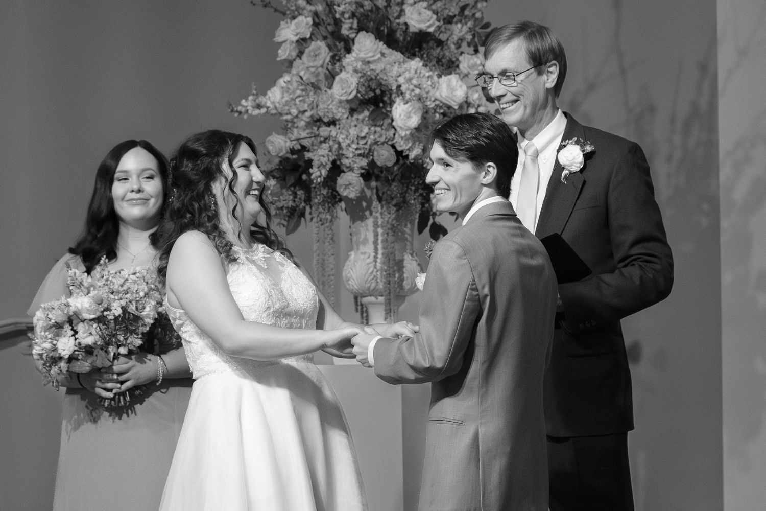 Black and white wedding ceremony photo of couple exchanging vows with bridesmaid and officiant.