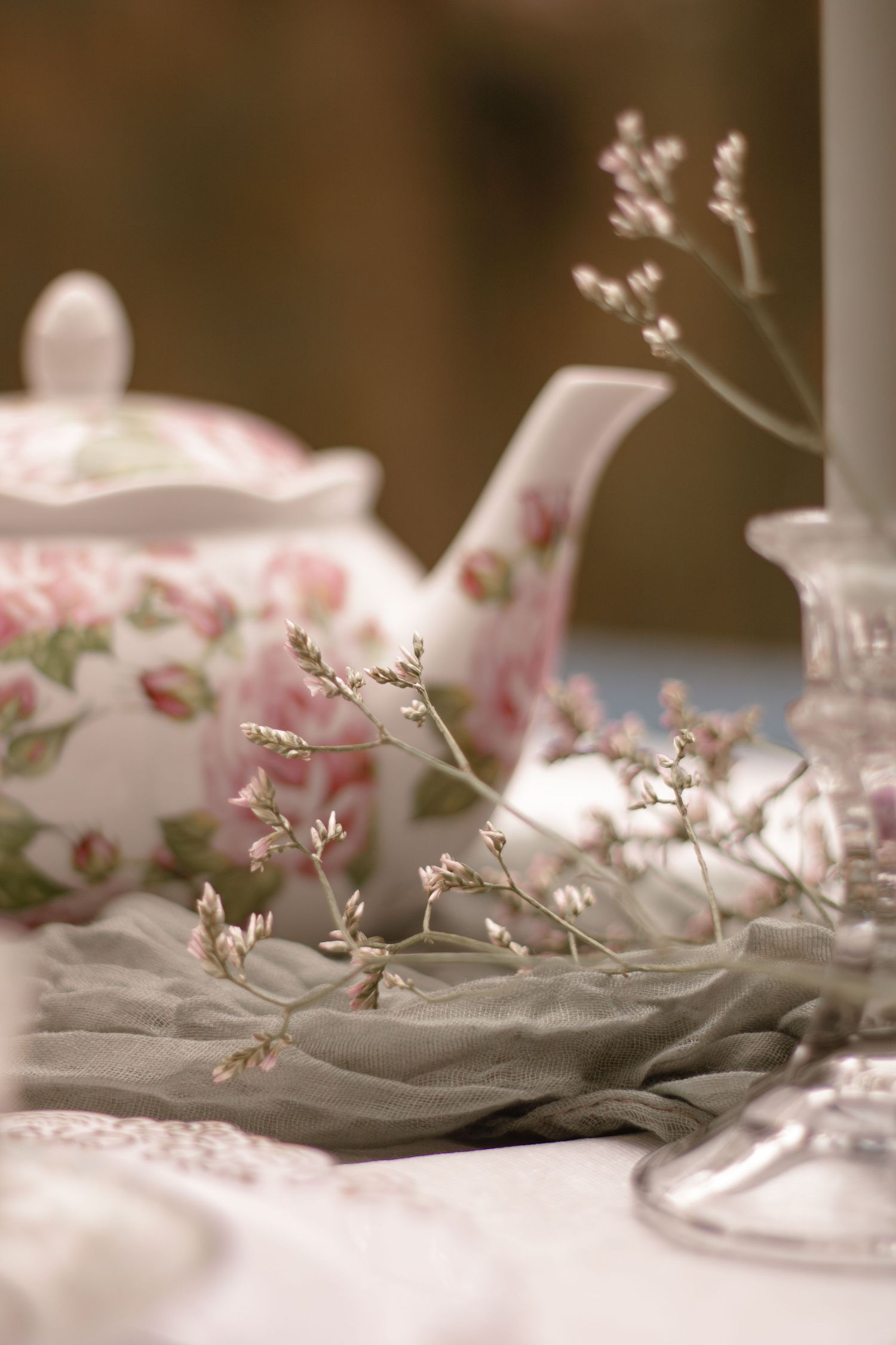 Delicate floral teapot with pink rose pattern sits beside wispy baby's breath flowers on soft gray fabric.