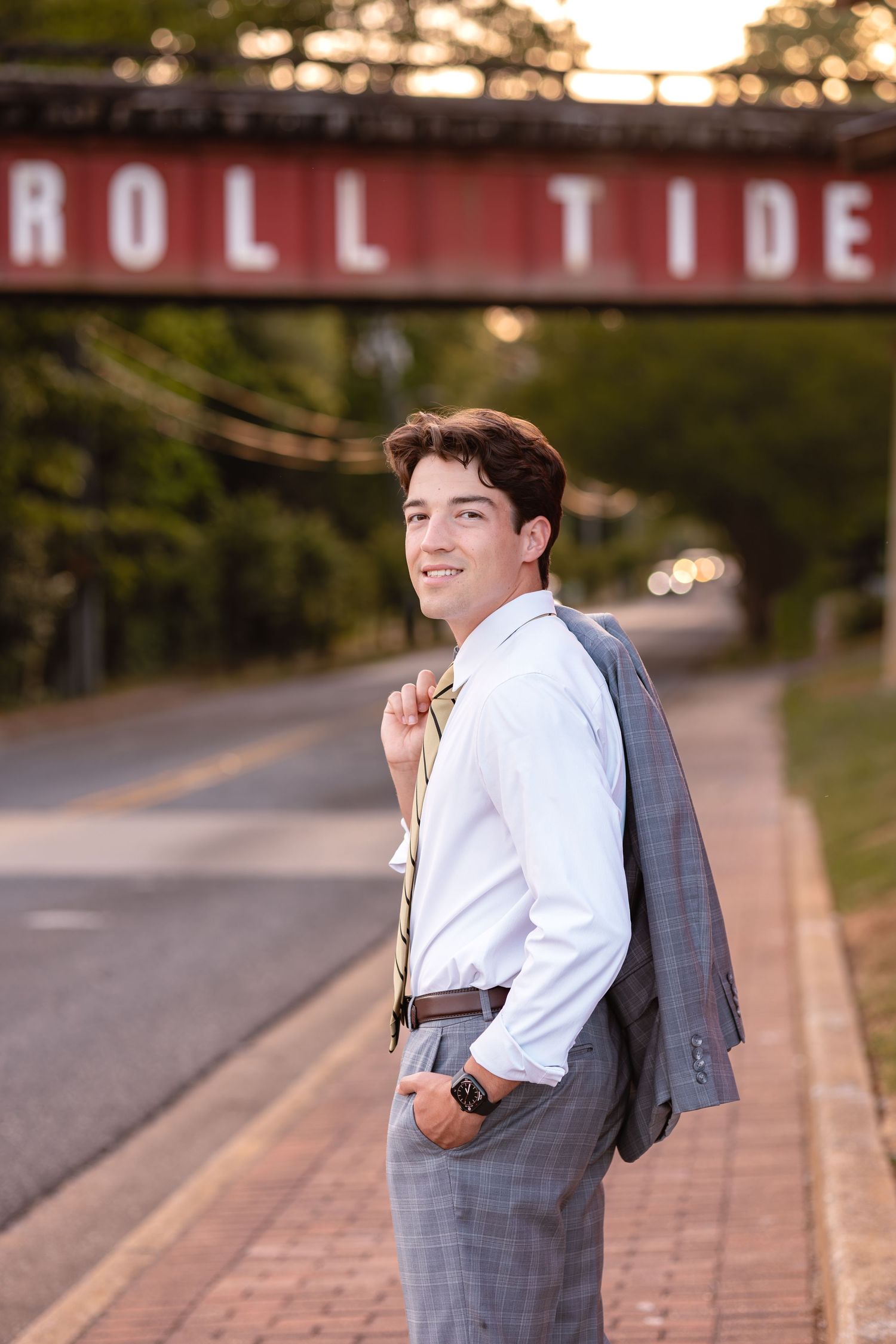 Young man in gray suit holding jacket over shoulder, smiling on a brick sidewalk near a Roll Tide bridge.