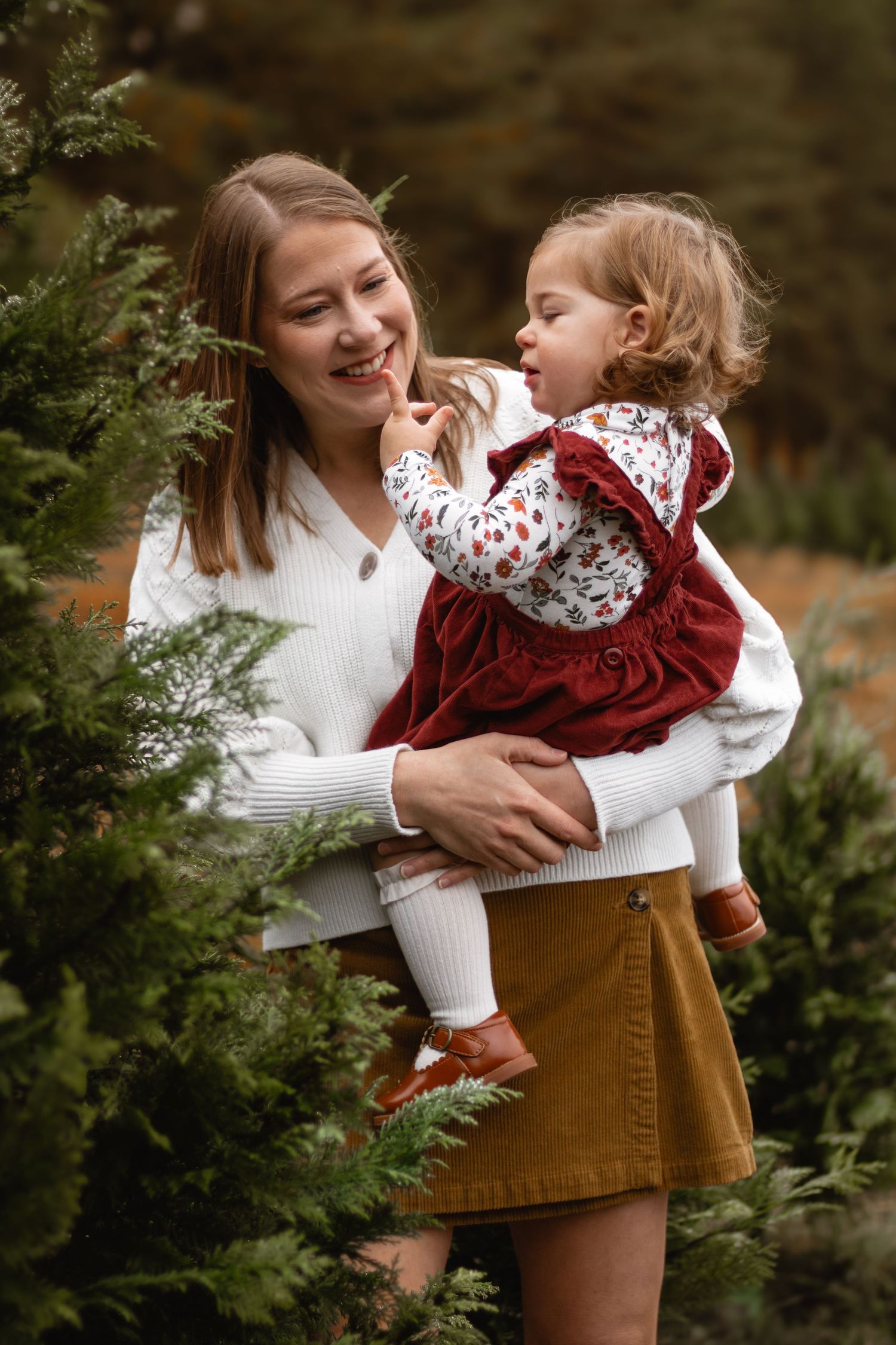 A smiling mother in a white sweater holds her toddler girl in a red dress among evergreen trees.