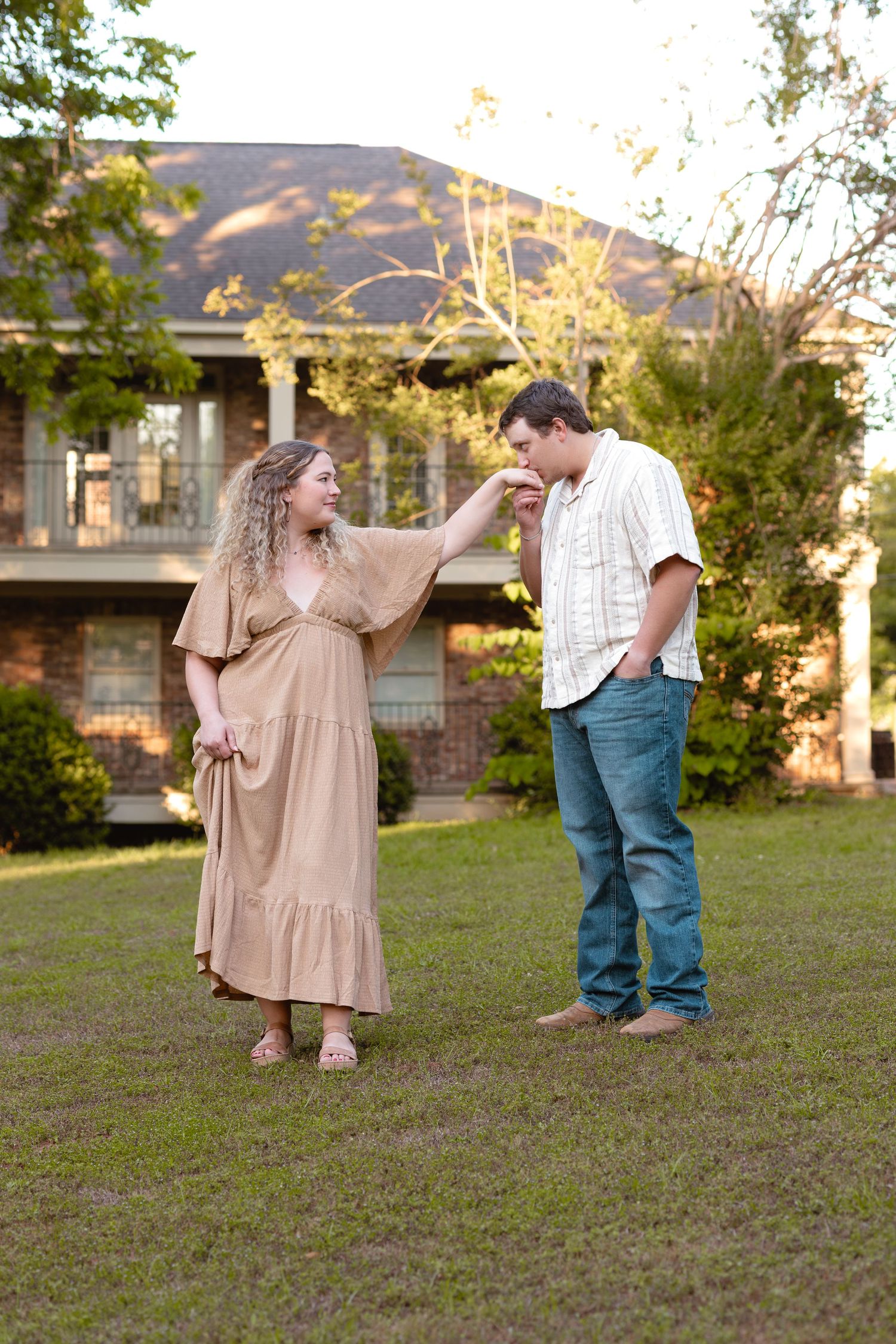 A couple shares a romantic moment outdoors, with the woman in a tan dress offering her hand to the man in jeans.