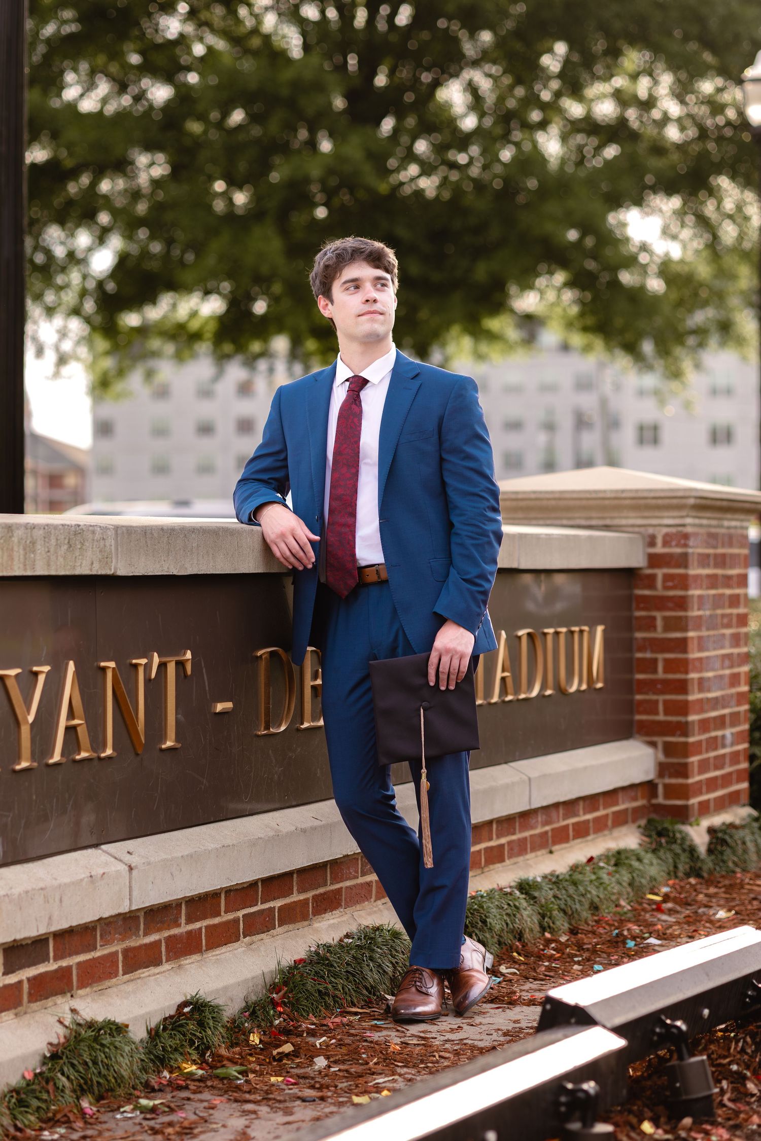 Young man in blue suit and red tie leaning against Bryant-Denny Stadium sign, holding a portfolio.