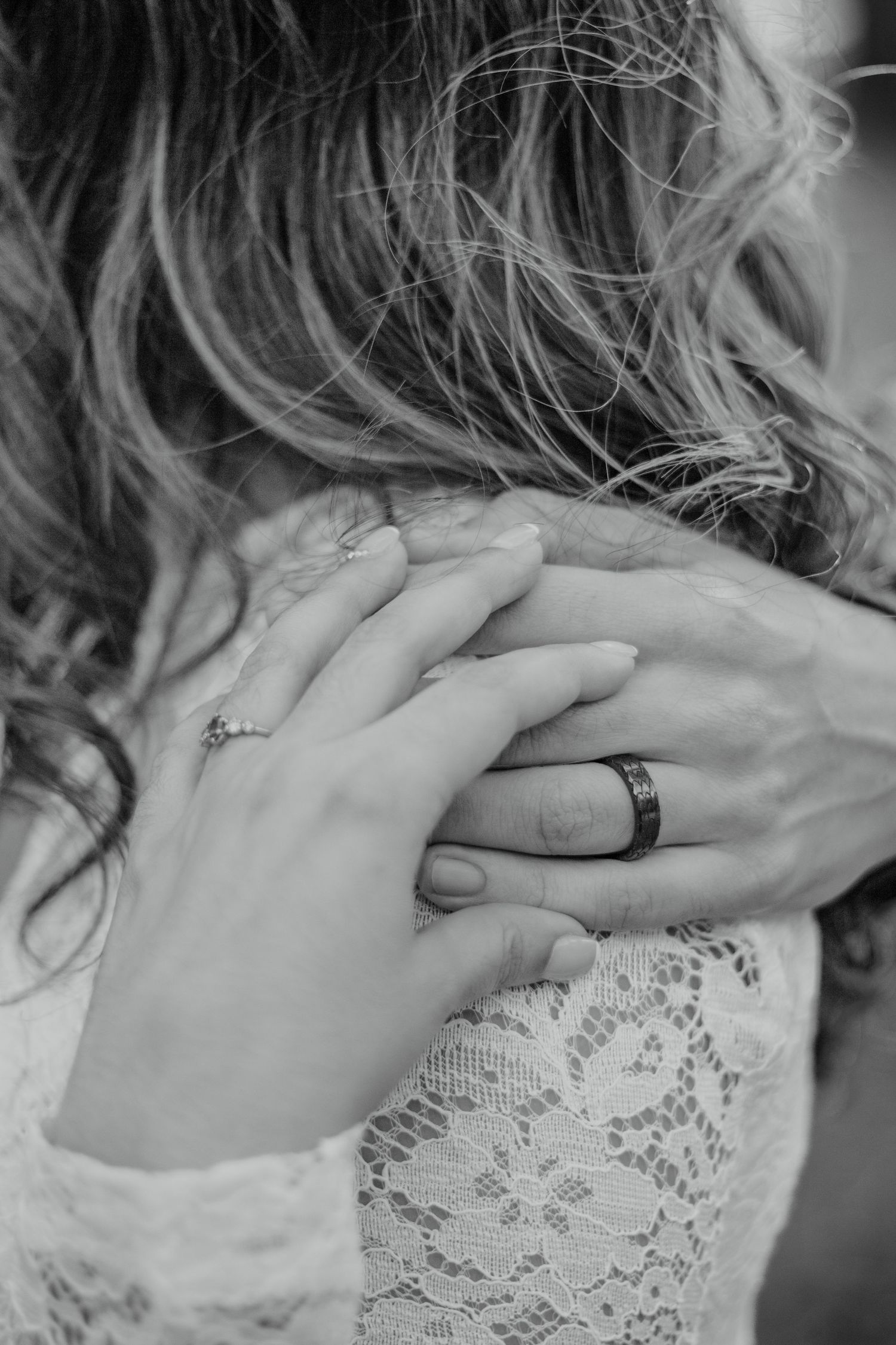 Close up black and white photo of hands with wedding rings against delicate white lace sleeve.