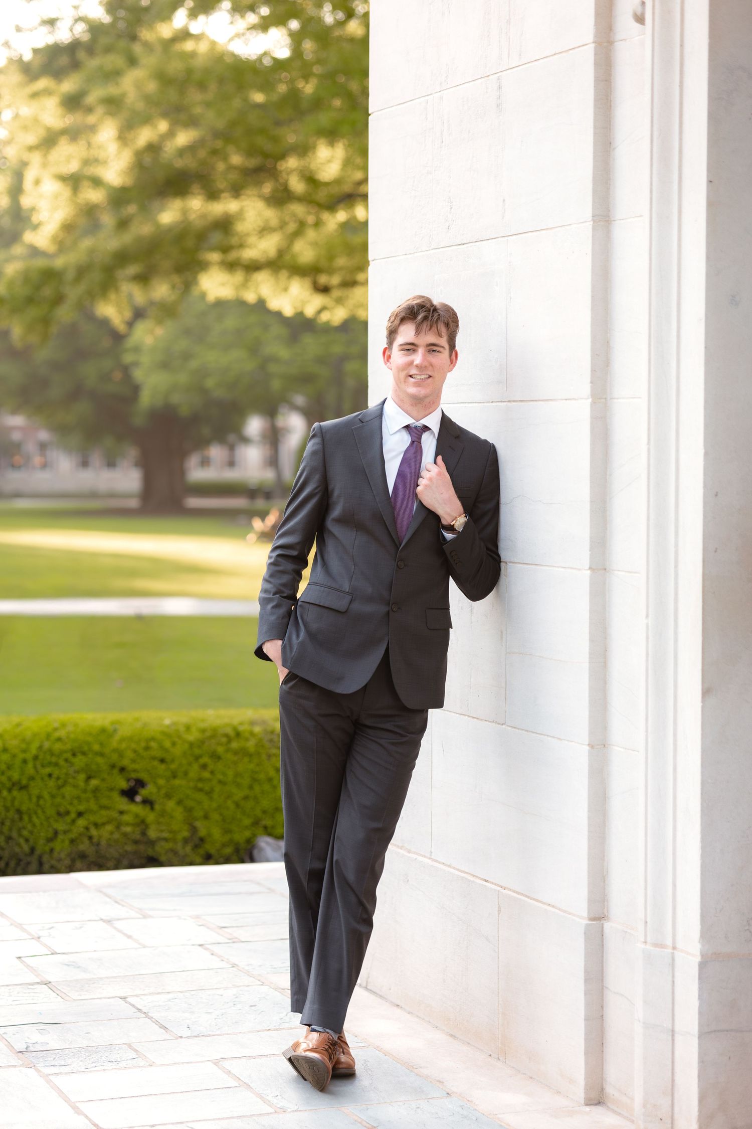 Young businessman in dark suit and purple tie leaning against white marble column in outdoor park setting.