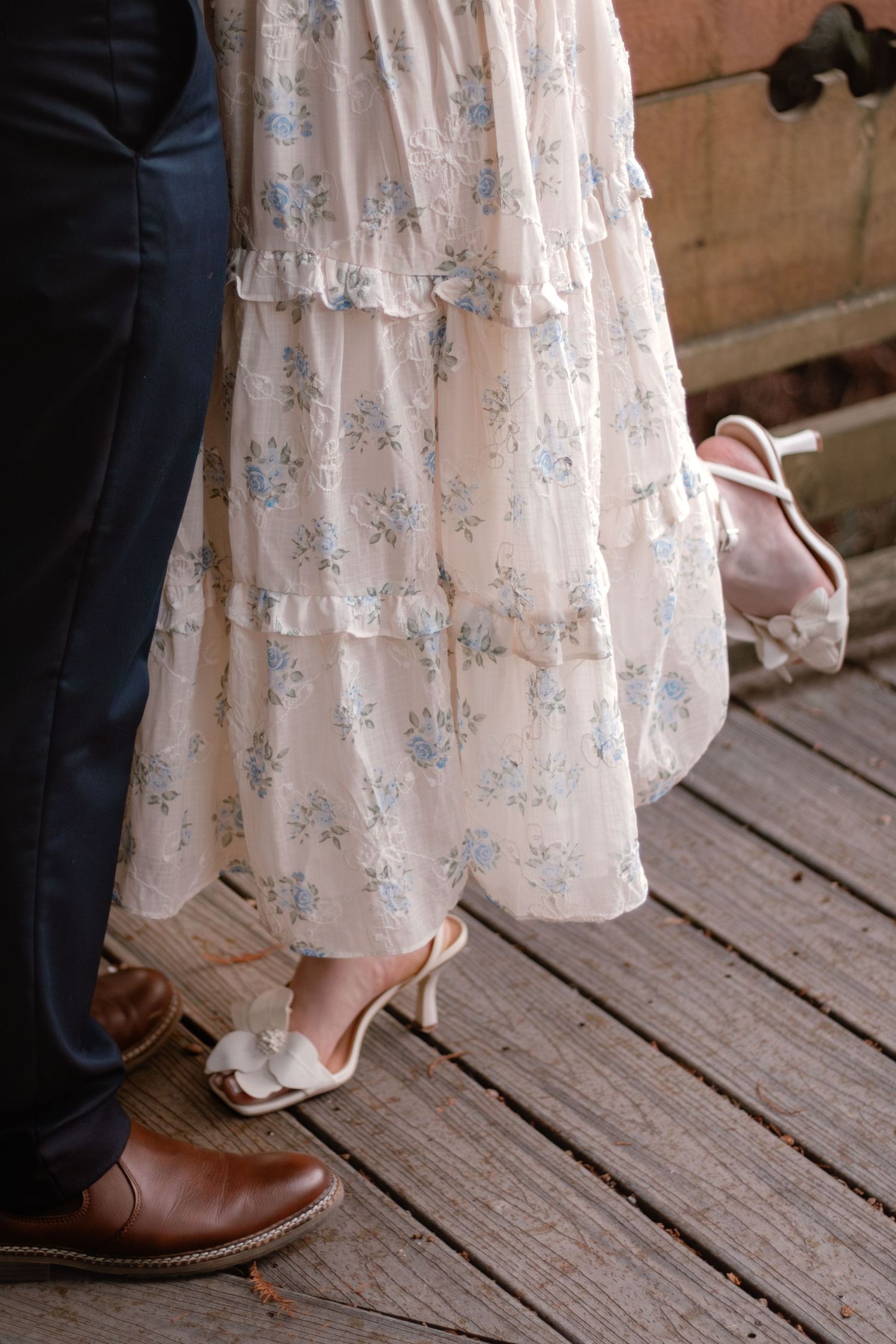 Couple standing on wooden deck, woman in floral tiered dress with heeled mules, man in navy trousers and brown leather shoes.