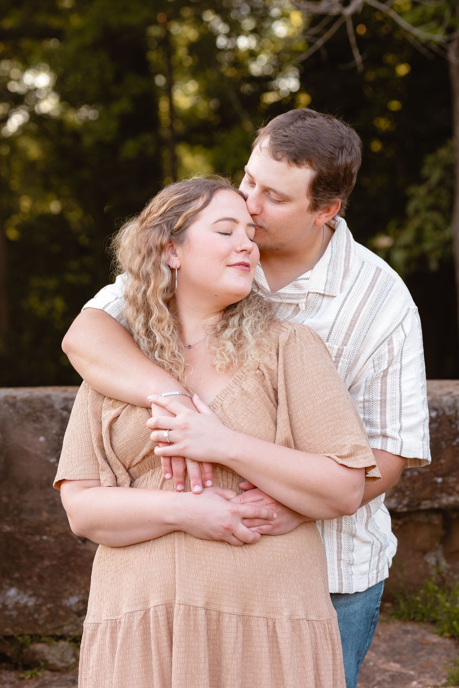 A man tenderly kisses a woman's cheek while embracing her from behind outdoors in a romantic couples portrait.
