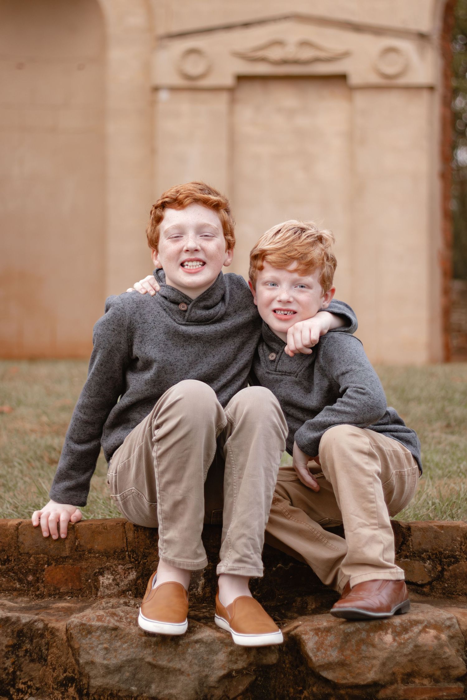 Two children sitting on a stone wall together sharing a laugh in gray sweaters and khaki pants.