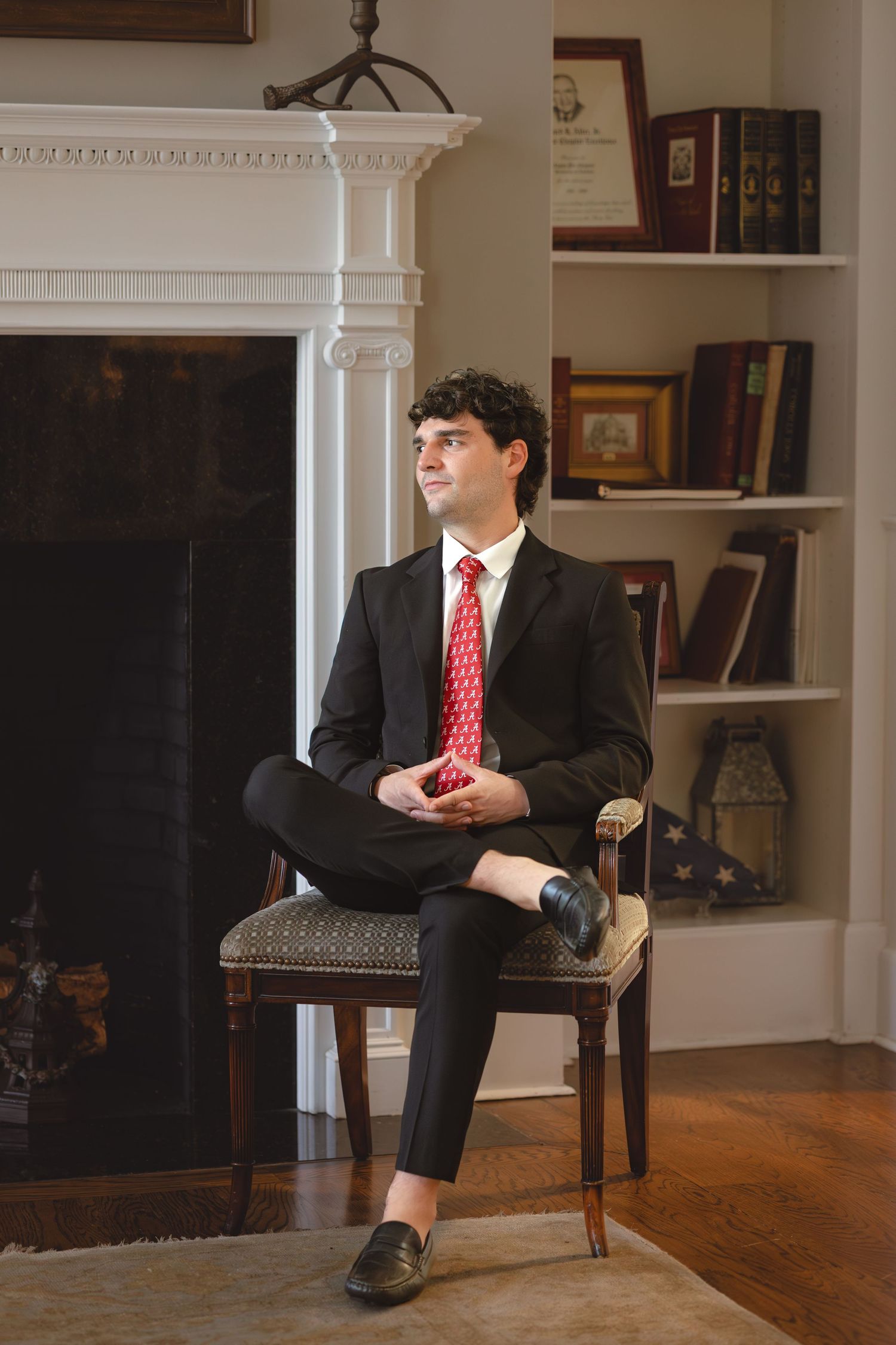 Young man in black suit and red tie sitting in wooden chair near fireplace with bookshelves in background.