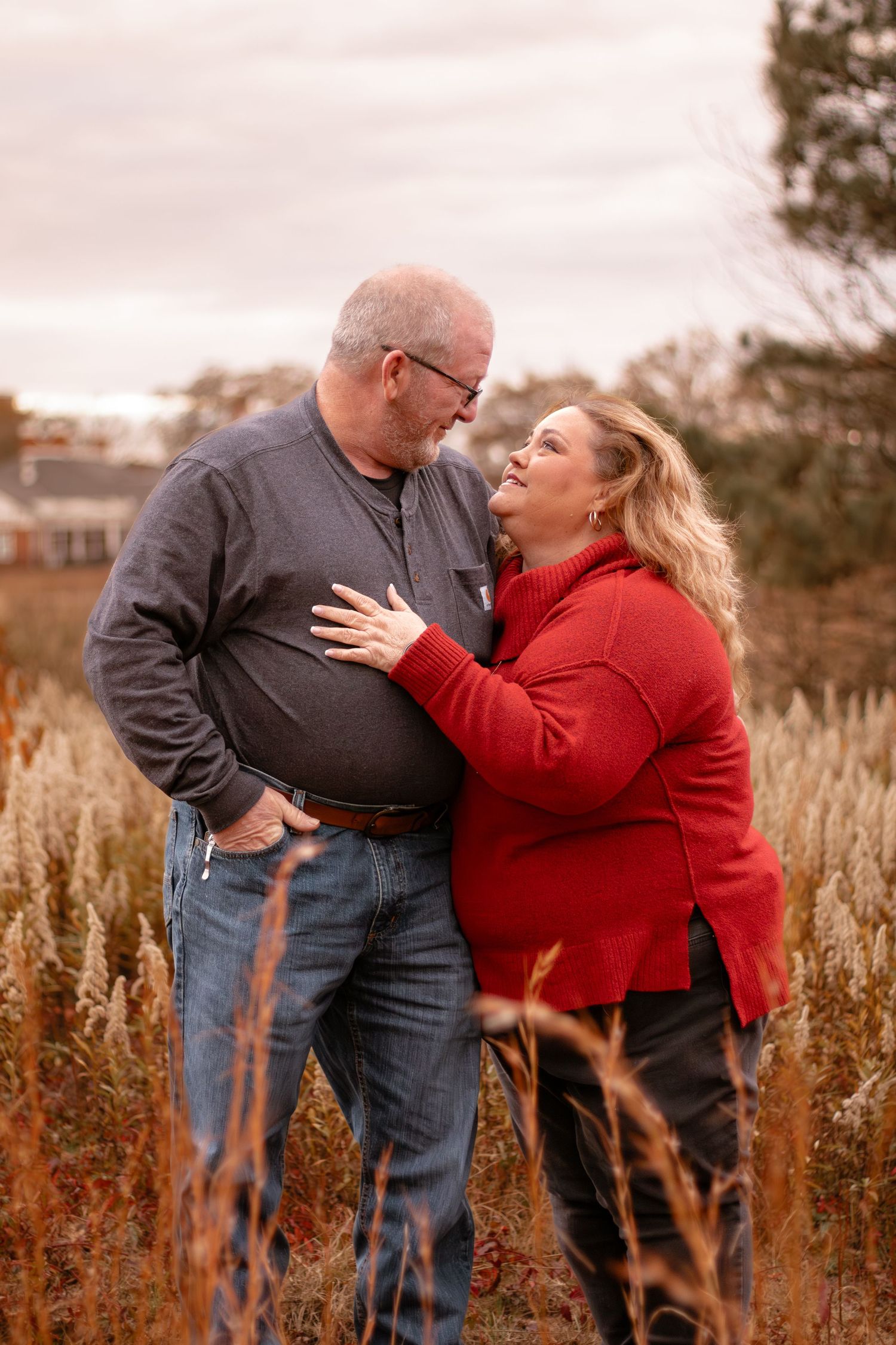 Two people embrace affectionately in an autumn field with tall brown grass at sunset.
