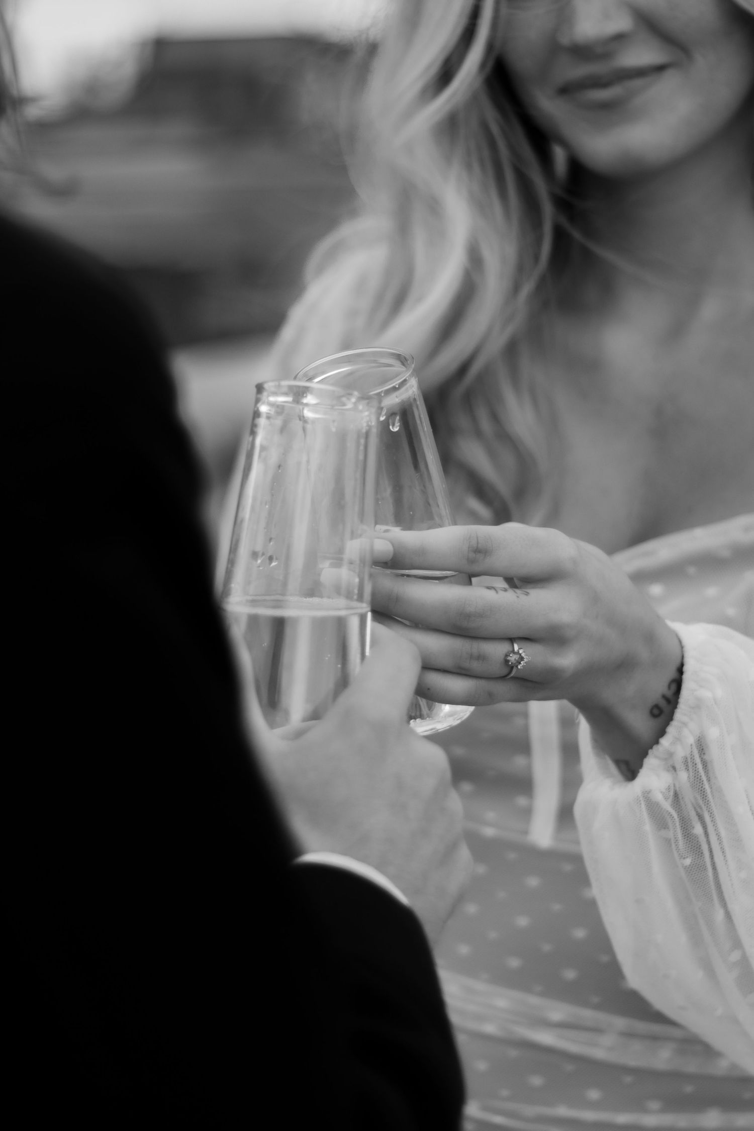 An elegant black and white shot of champagne glasses clinking in a toast with a diamond ring visible on one hand.