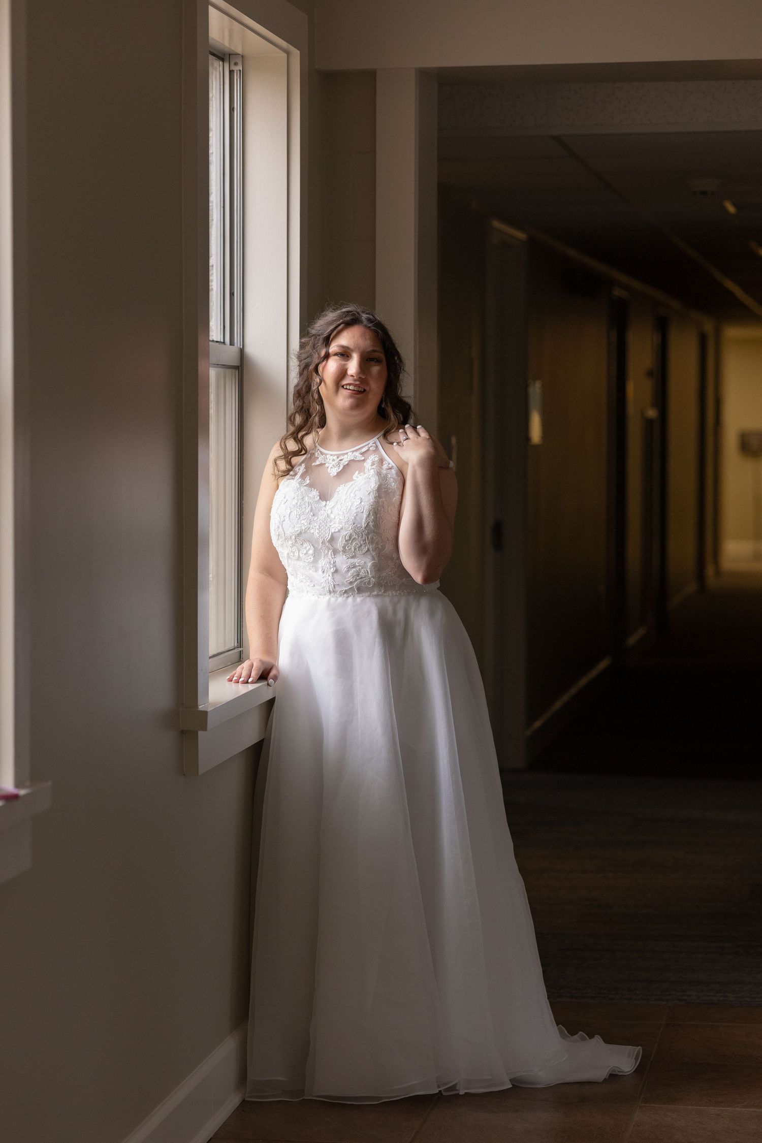 Bride in white lace ballgown wedding dress standing by window in elegant indoor hallway, soft natural light.