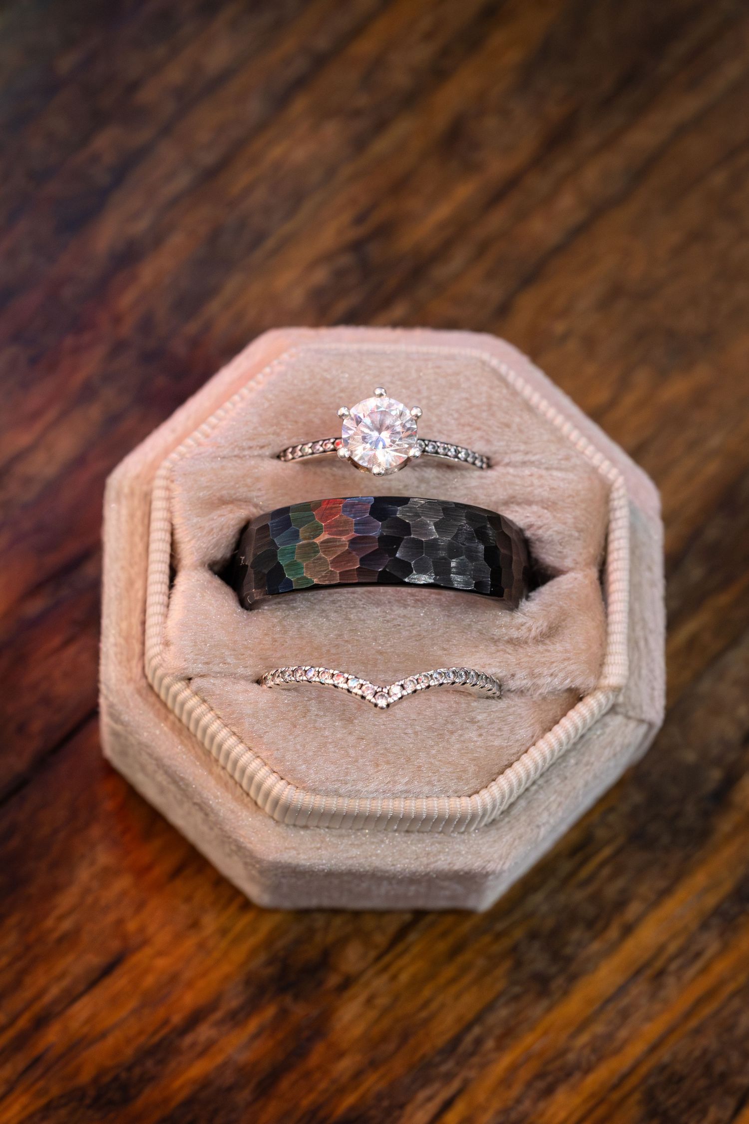 Three wedding rings including a diamond engagement ring and black band displayed in a beige octagonal ring box.