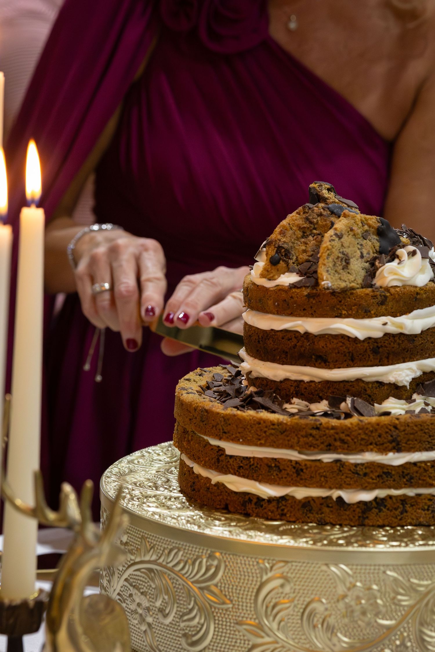 Woman in burgundy dress cutting a rustic naked layered wedding cake on gold stand with candles.