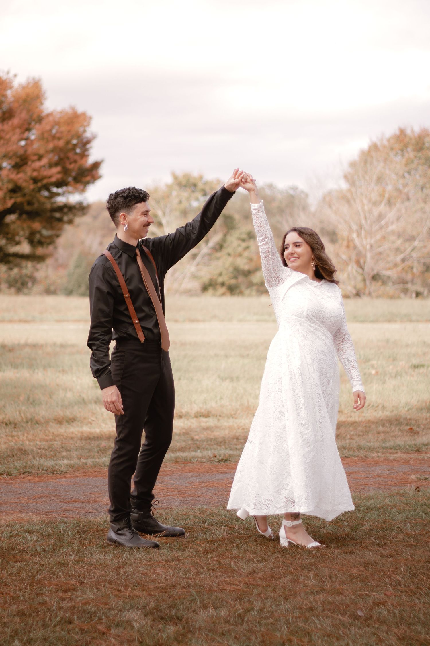 A bride and groom dance outdoors in a fall setting, she in white lace dress, he in black suit with brown suspenders.