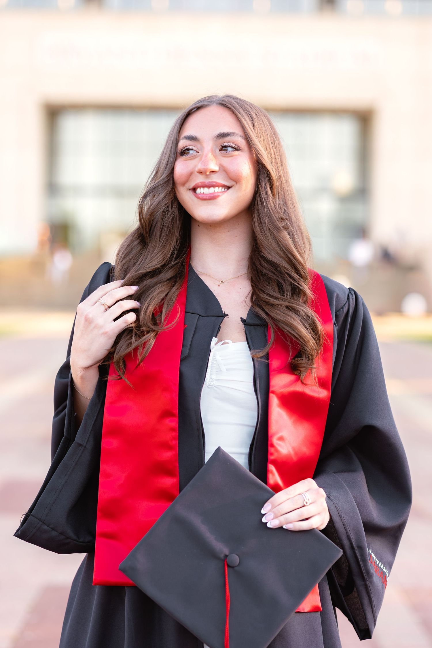 Smiling young woman in black graduation gown with red stole holding cap outdoors on graduation day.