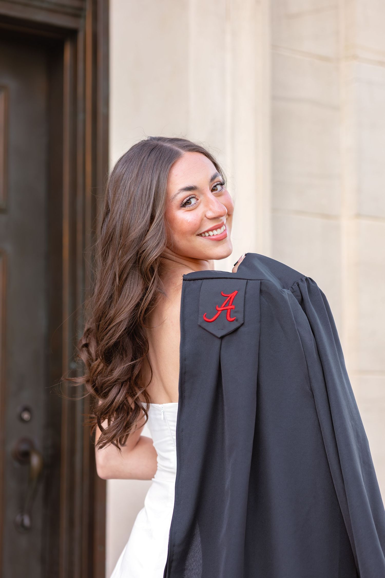 Smiling young woman holding a black University of Alabama graduation gown over her shoulder, wearing a white dress.