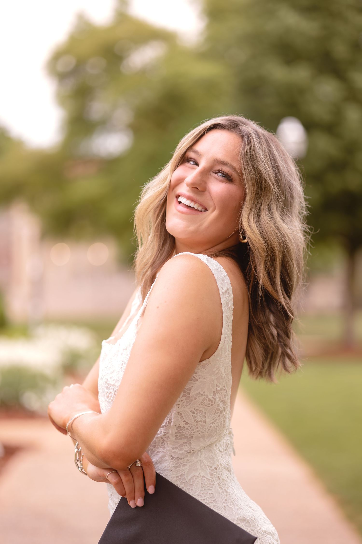 Smiling young woman in white lace top poses outdoors on a tree-lined path in a professional portrait photo.