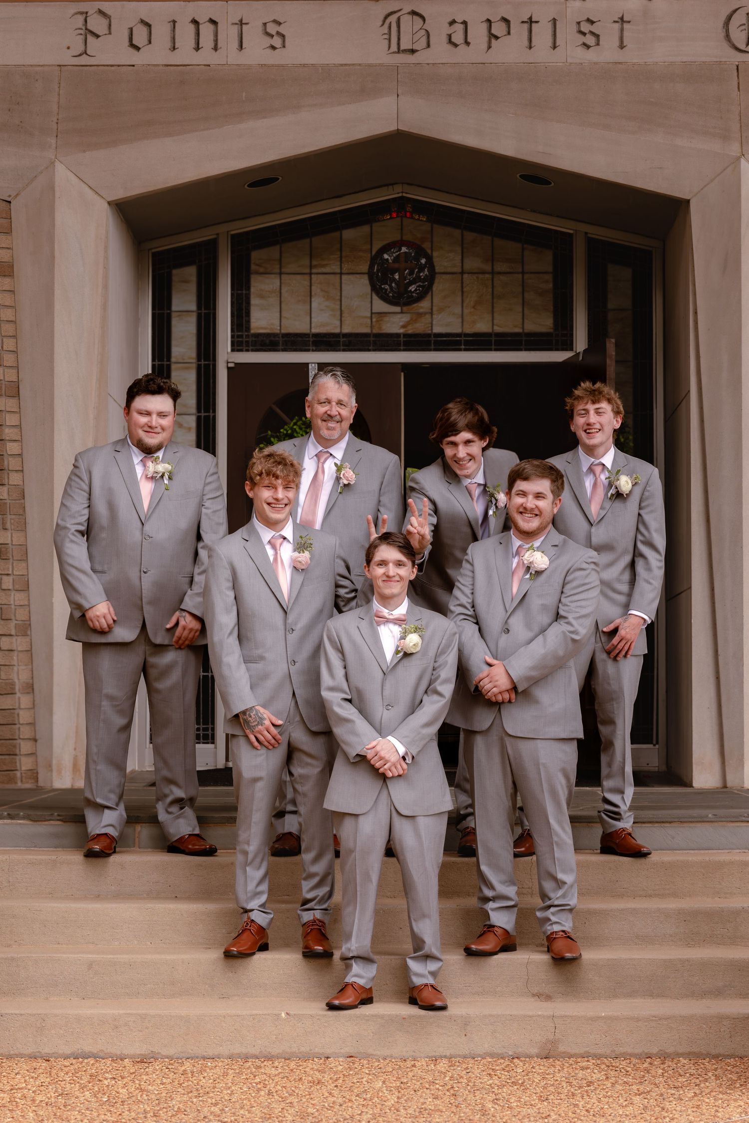Groom and groomsmen in gray suits with pink boutonnieres pose on church steps for a classic wedding party photo.