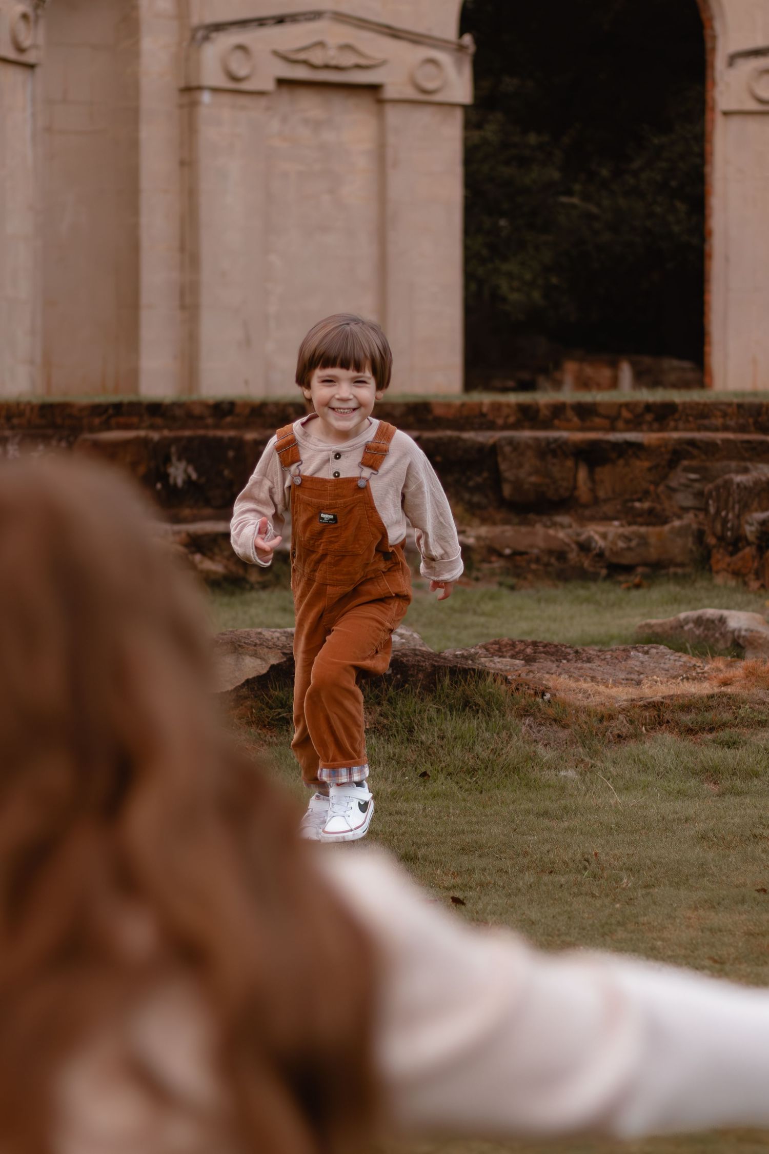 A child in orange overalls runs playfully in front of a classical architectural structure.
