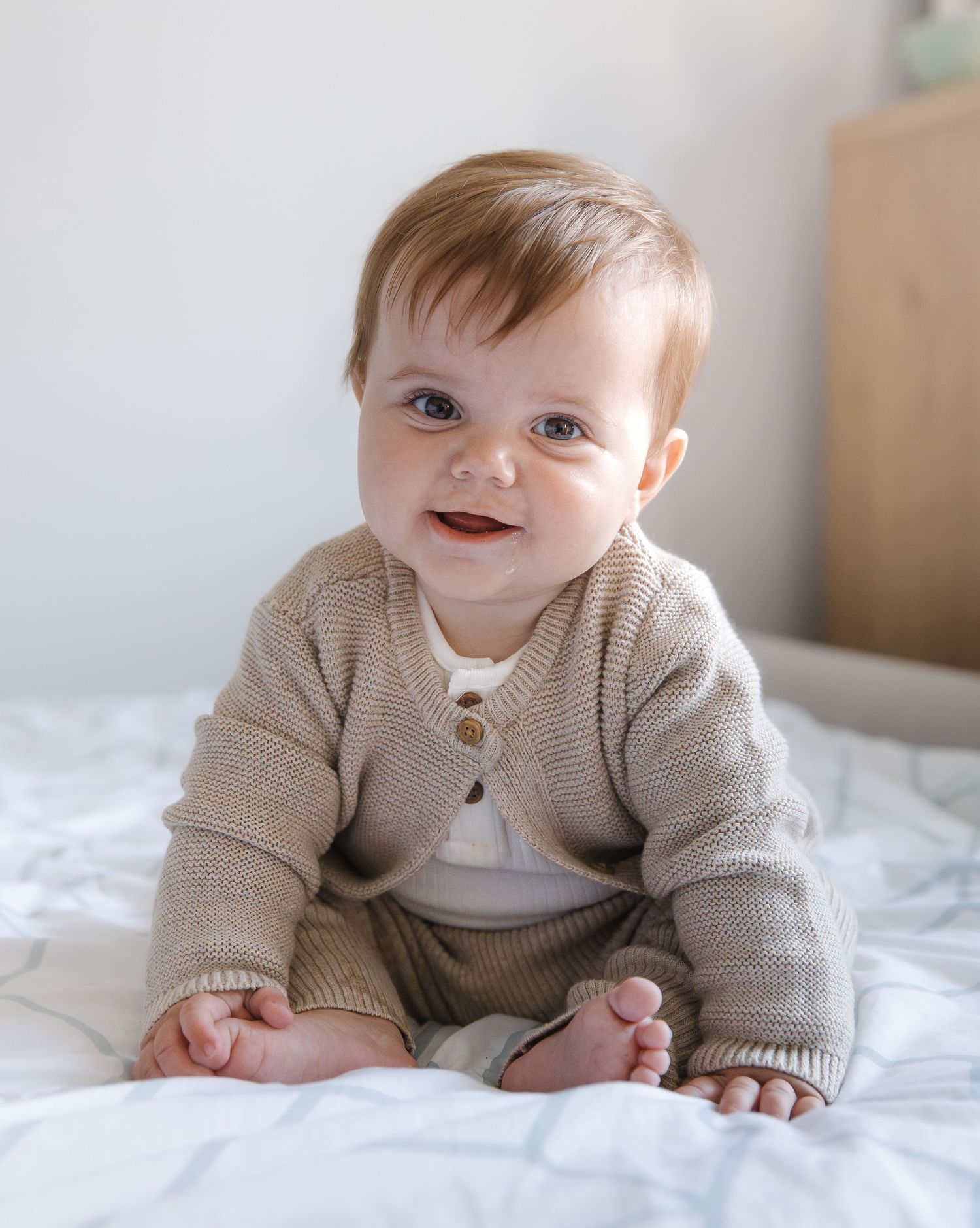 A baby in a beige knit sweater lies on white bedding while smiling and playing.