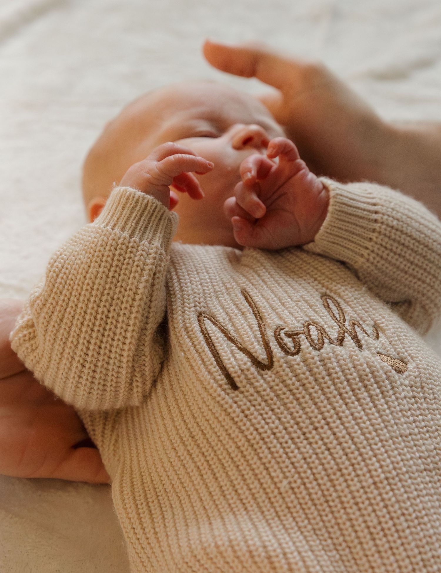 A tiny baby wearing a cozy beige knit sweater with 'Noah' text stretches while lying on soft fabric.