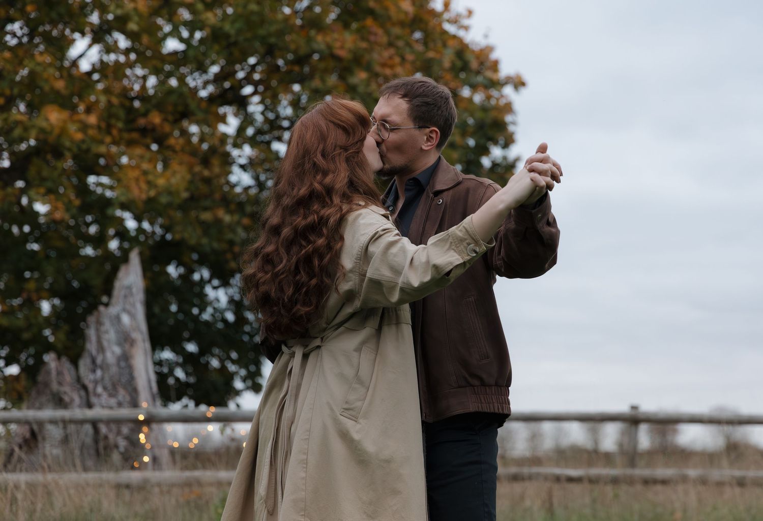A couple in autumn attire dances together in front of fall foliage at an outdoor location.