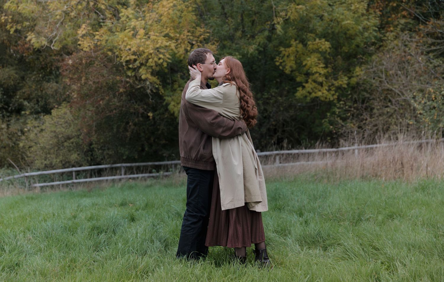 A couple embraces in a tender moment on a grassy hill with autumn trees in the background.
