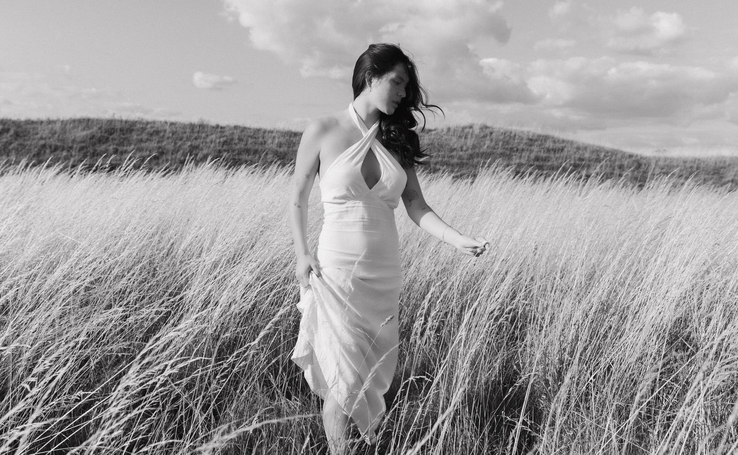 Ethereal figure in white dress moves through tall grass field under cloudy sky.