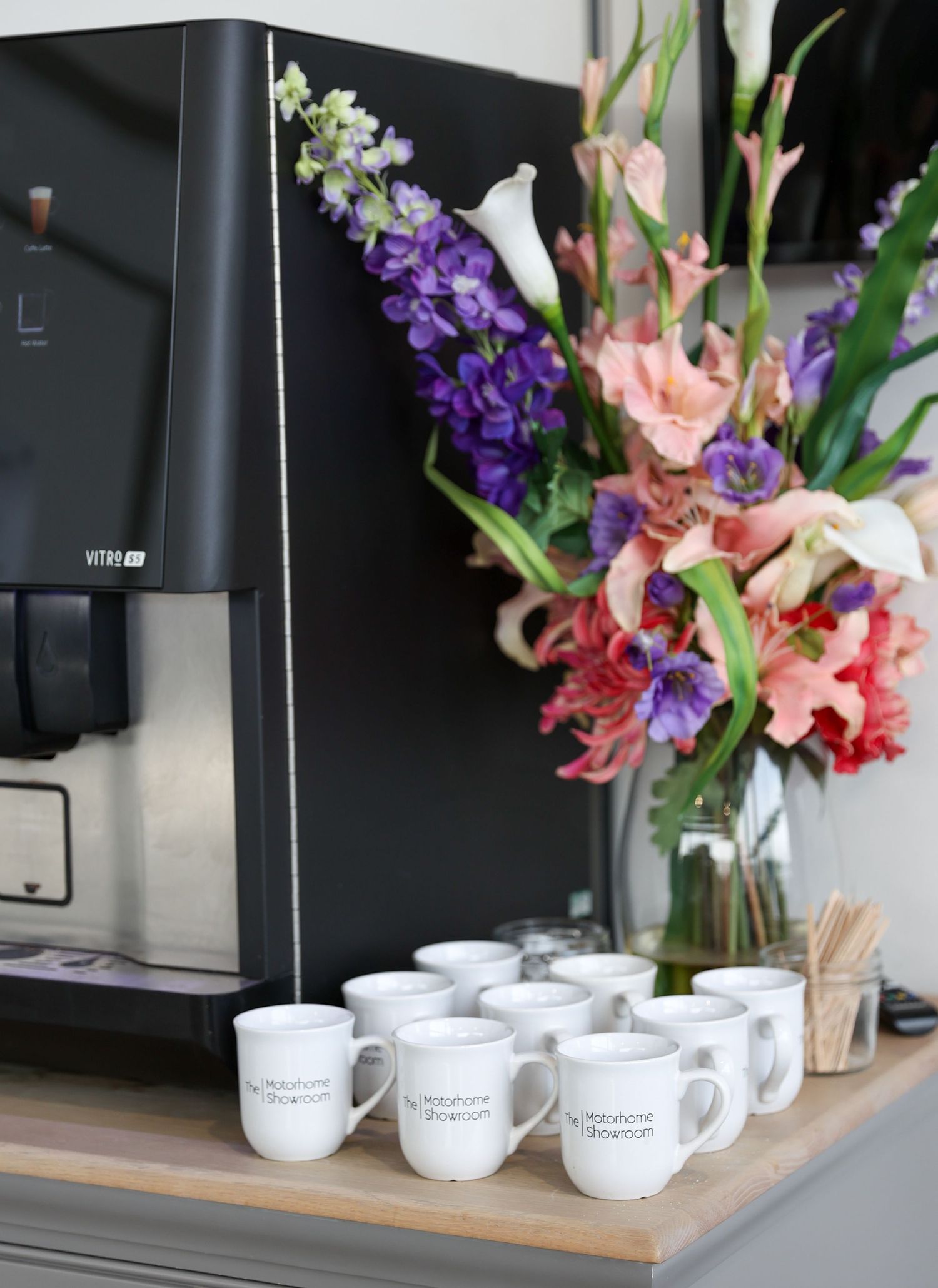 White coffee mugs arranged on wooden tray with colorful floral arrangement in background.