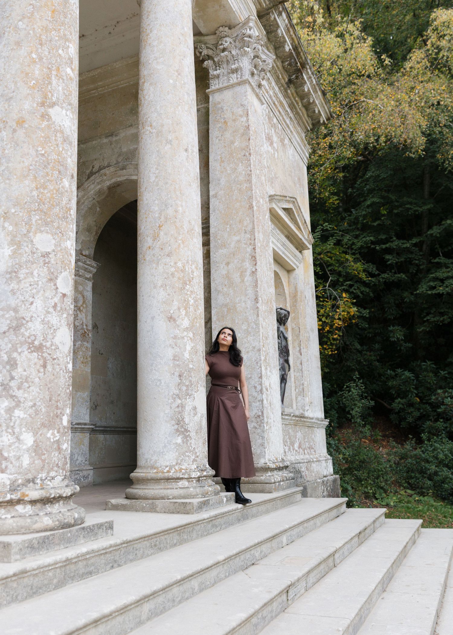 A figure stands in a brown dress at the entrance of a classical stone building with white columns and stone steps.