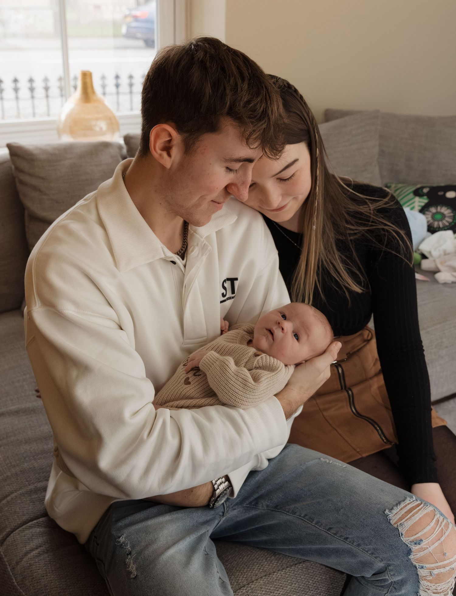 A couple in casual attire sits close together on a couch, sharing an intimate moment with a small infant.