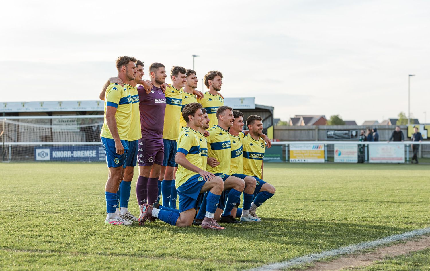 Football team posing for a group photo on a grass pitch in yellow and blue kits before a match.