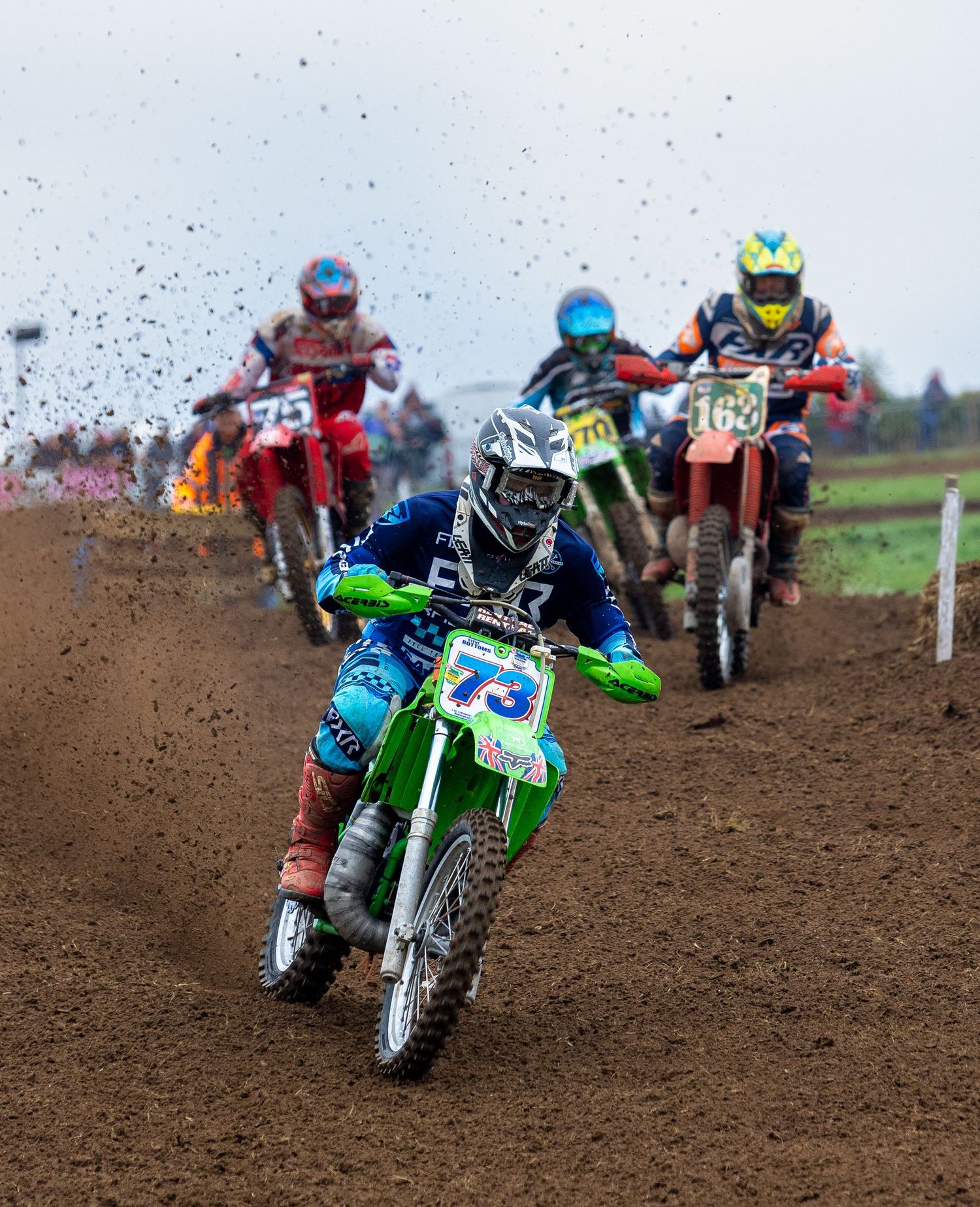 Motocross riders racing through a dirt track kicking up mud and dirt as they navigate the course in intense competition.