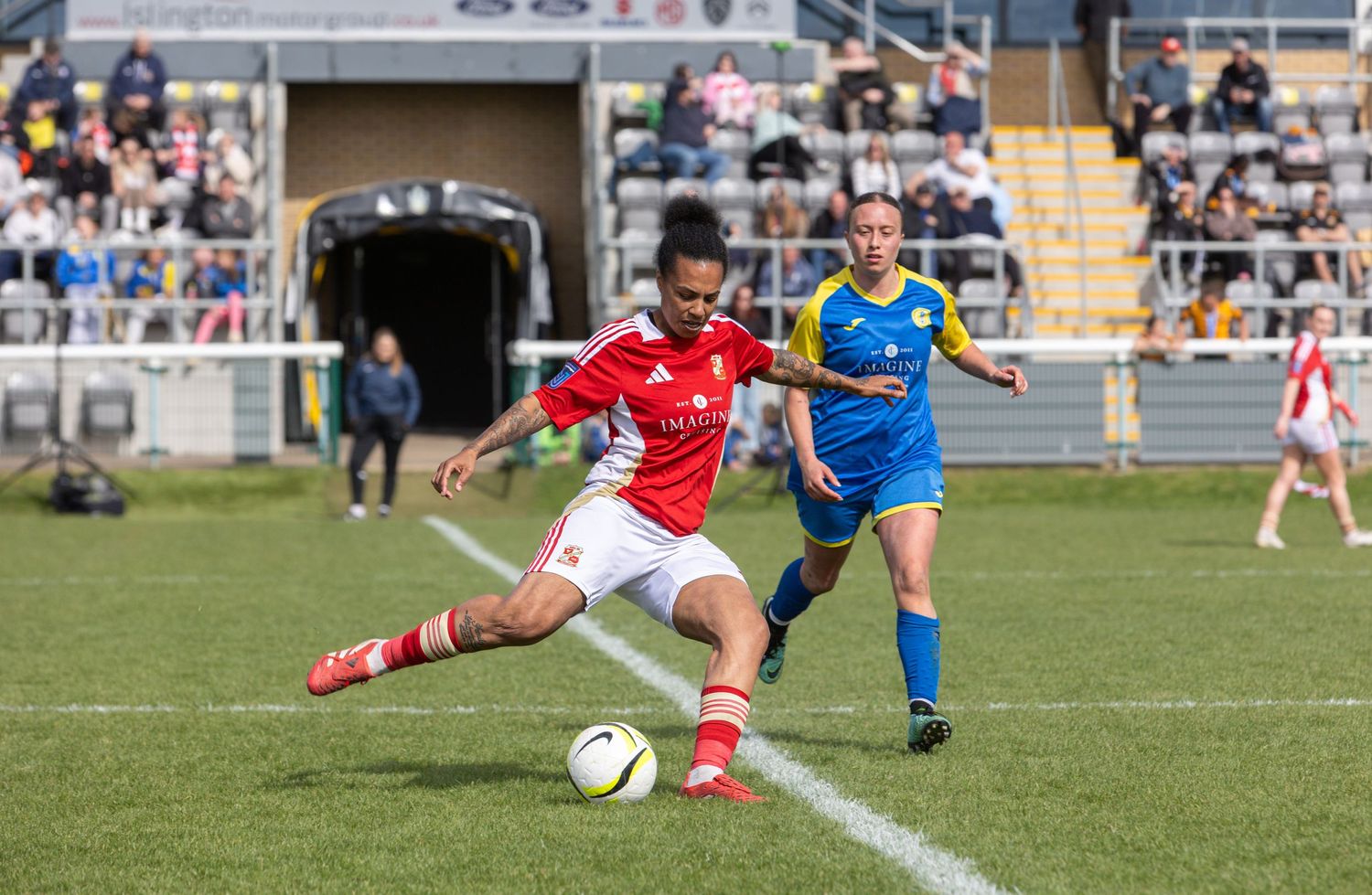 Two women soccer players compete on a grass field, one in red and white, the other in blue and yellow kit.